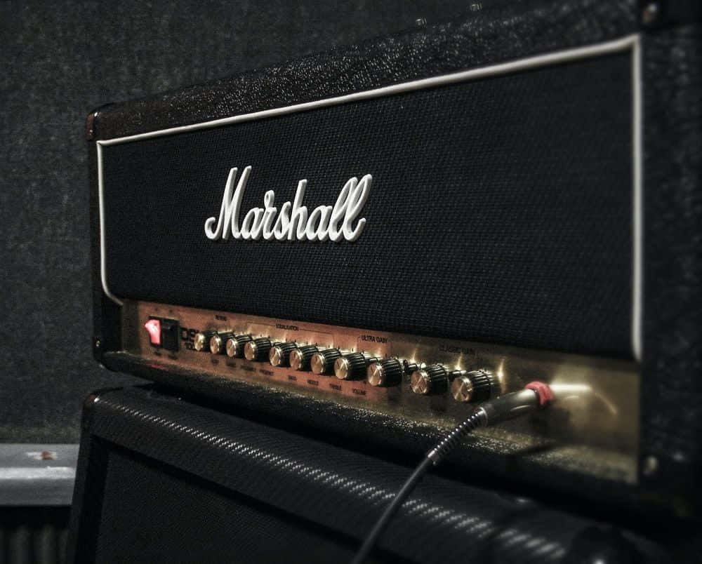 電吉他靜置於擴音器前，準備登台演出 壁紙 — 復古黑色Marshall結他擴音器特寫，帶有發光旋鈕和紅色電源指示燈，背景為深色紋理牆壁。