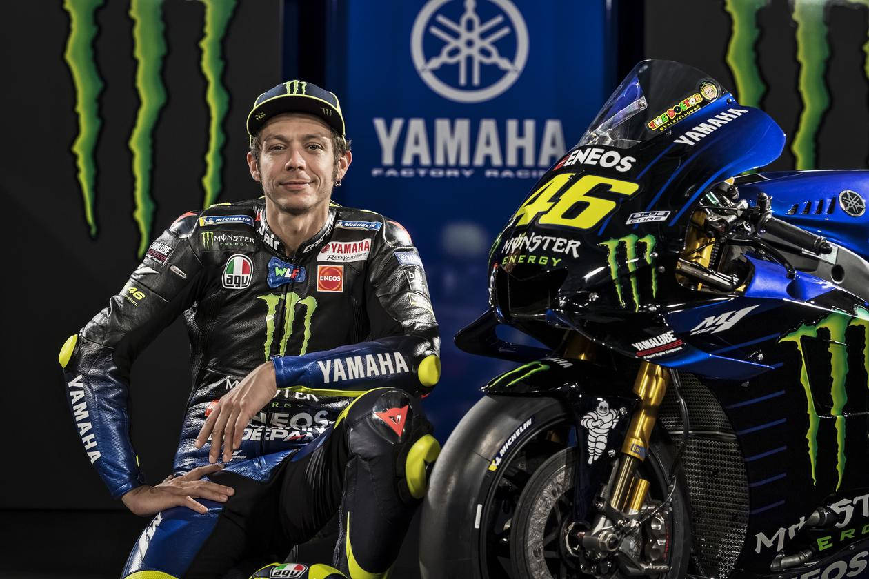 Monster Energy 桌布 壁紙 — 電單車賽車手華倫天奴·羅西自信地擺姿勢，站在他飾有Monster Energy標誌的藍黑色Yamaha MotoGP電單車旁。