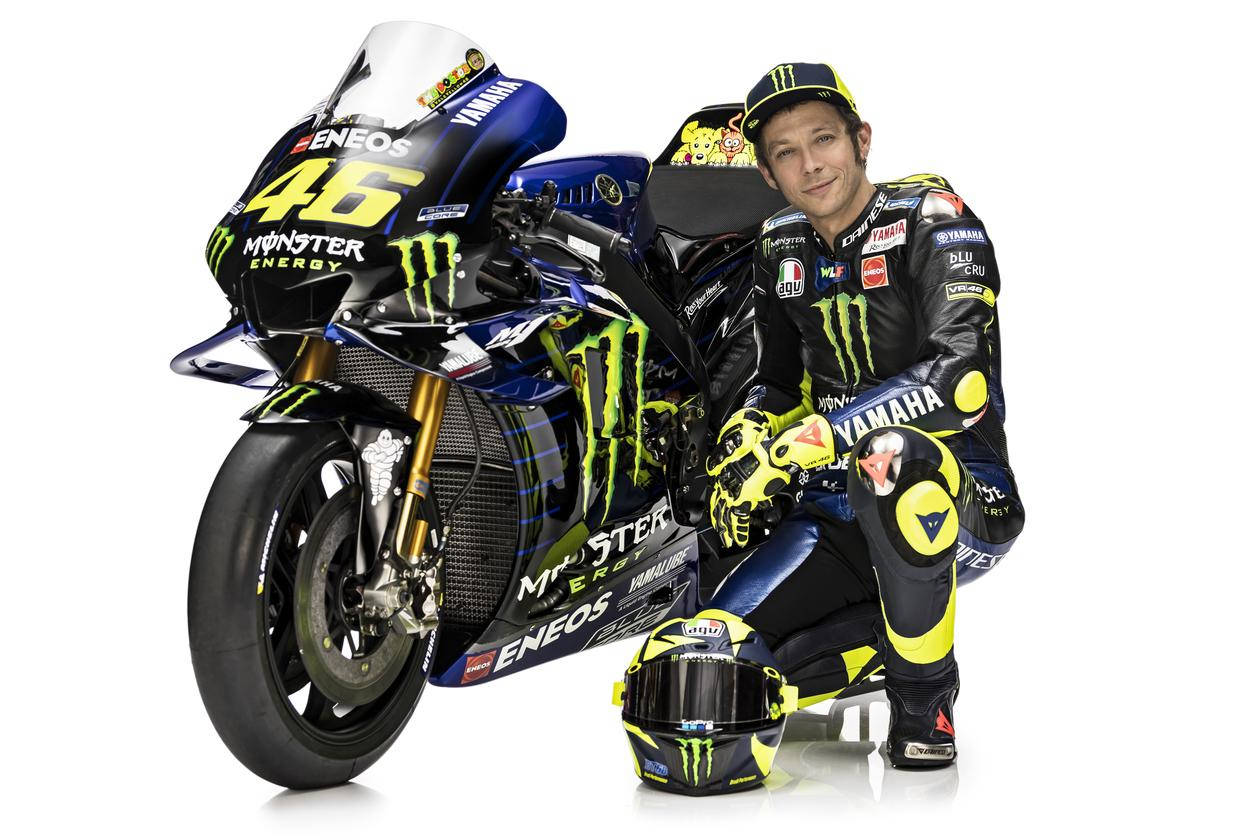 Monster Energy 桌布 壁紙 — 電單車賽車手華倫天奴·羅西身穿全套裝備，跪在他藍黃色的Yamaha MotoGP電單車旁，背景為白色。
