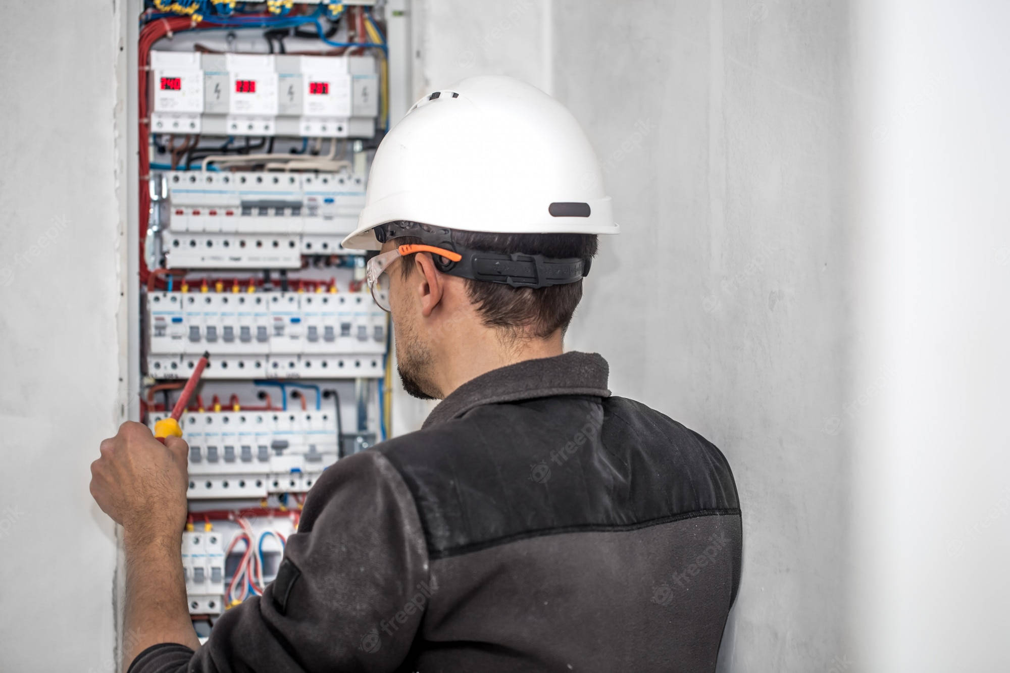 電工檢查電壓 壁紙 — 戴白色頭盔和安全眼鏡的電工正在安裝牆上電箱，內有彩色電線和斷路器。