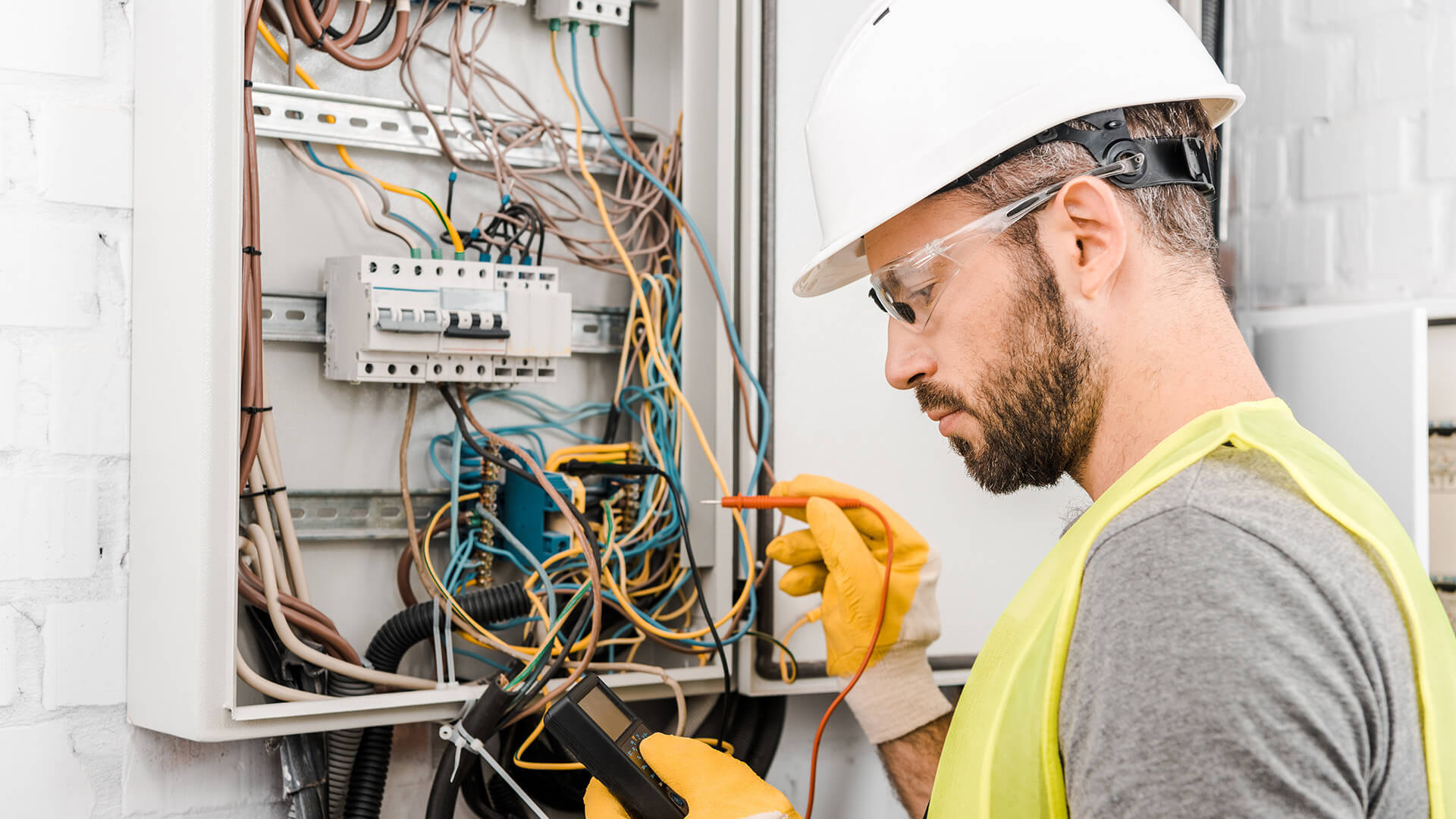 電工檢查電線 壁紙 — 電工戴著頭盔和安全背心，用萬用電錶測試打開的電箱內的接線。
