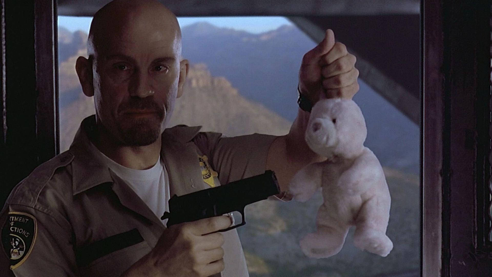 電影《Con Air》中的反派角色Cyrus 'The Virus' Grissom 壁紙 — 一個光頭男人穿著警長制服，在群山背景下拿著一把槍和一隻白色泰迪熊。