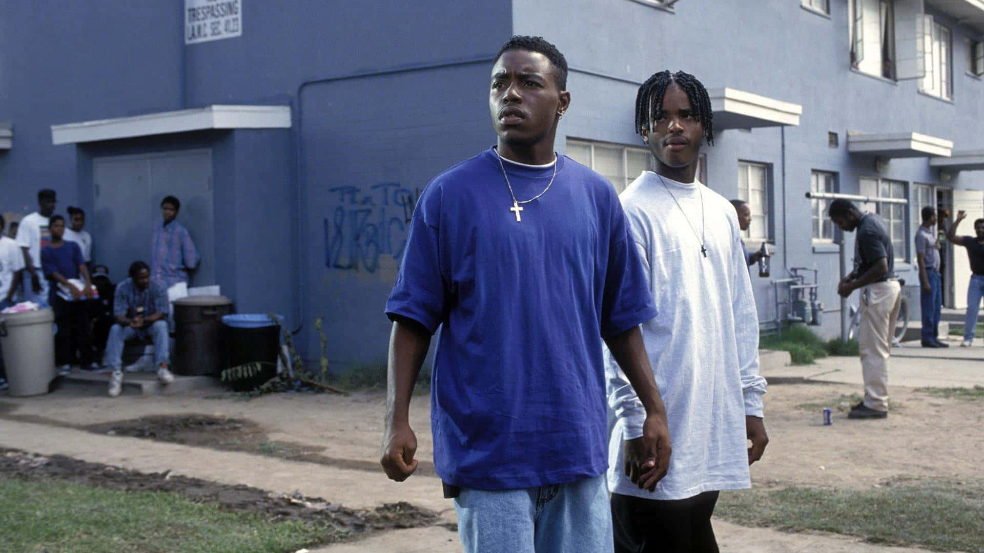 電影《Menace II Society》主角卡恩與O-Dog 壁紙 — 兩名身穿藍白襯衫的年輕男子表情嚴肅地向前走，經過一棟帶有塗鴉的藍色公寓大樓。