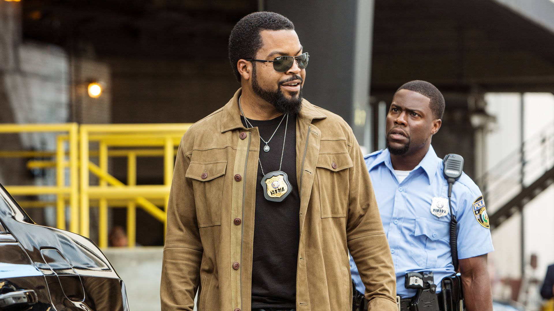 電影《Ride Along》中的Ice Cube 壁紙 — 兩名男子，一人身穿淺褐色外套並佩戴徽章，另一人身穿警察制服，站在工業環境中的一輛黑色汽車旁。