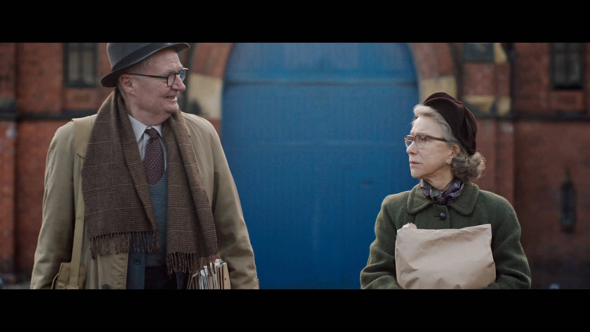 電影《The Duke》劇照：吉姆·布勞德本特飾演Kempton Bunton 壁紙 — 兩位穿著復古外套和帽子的長者，在一條磚街上，旁邊有一扇大藍門，親切交談。
