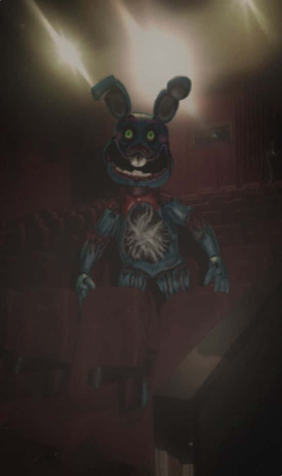電影院中的恐懼邦妮 FNAF 圖片 壁紙 — 一隻帶有發光綠色眼睛的威脅性藍色兔子機械動畫坐在昏暗的劇院中，聚光燈照射下來。