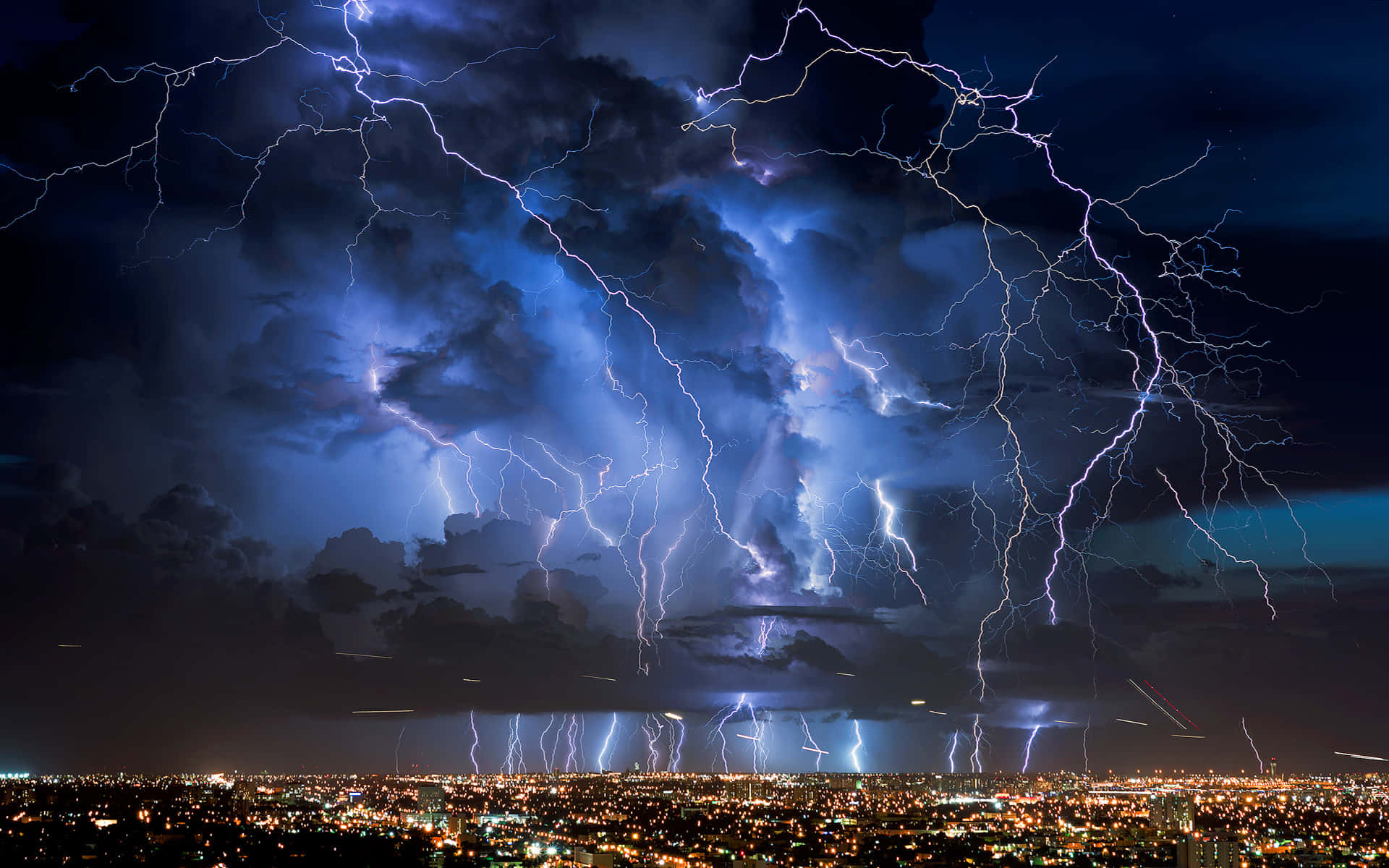 電能填滿夜空 壁紙 — 強烈的閃電照亮了暴風雨夜空中廣闊的城市景觀，城市景觀閃爍著人造燈光。