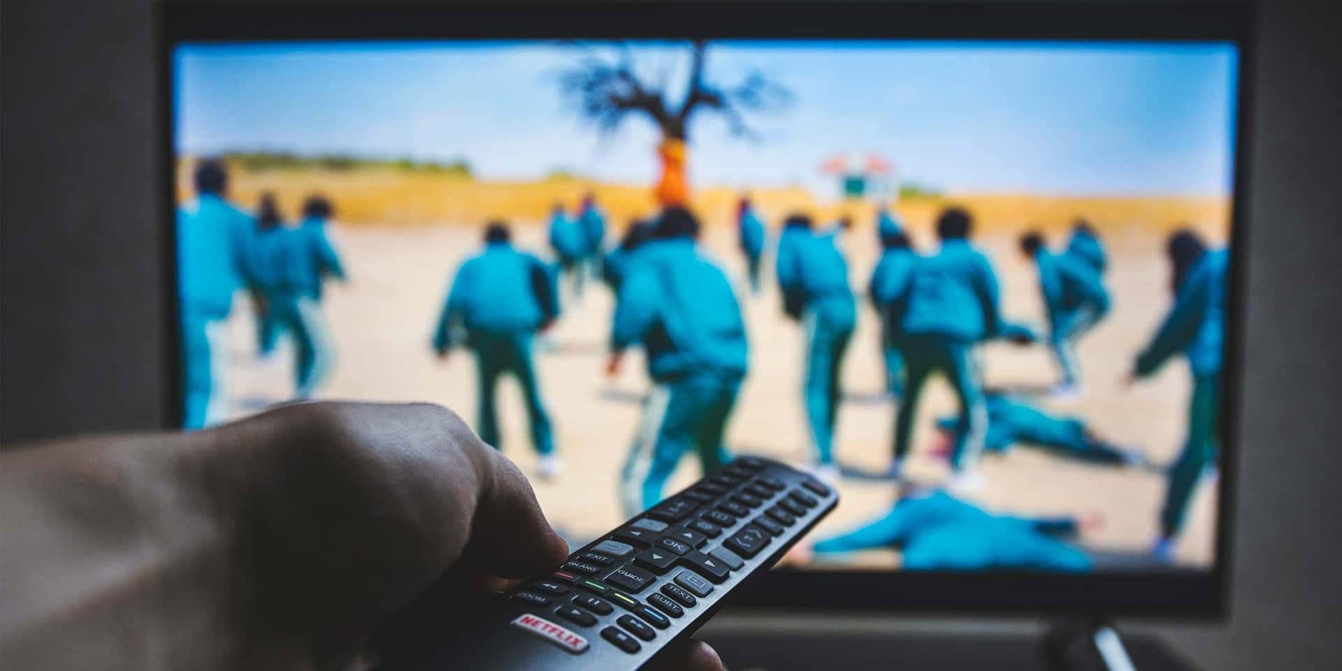 手拿 Netflix 遙控器指向電視屏幕，屏幕顯示沙漠景觀中穿著藍色制服的人們。