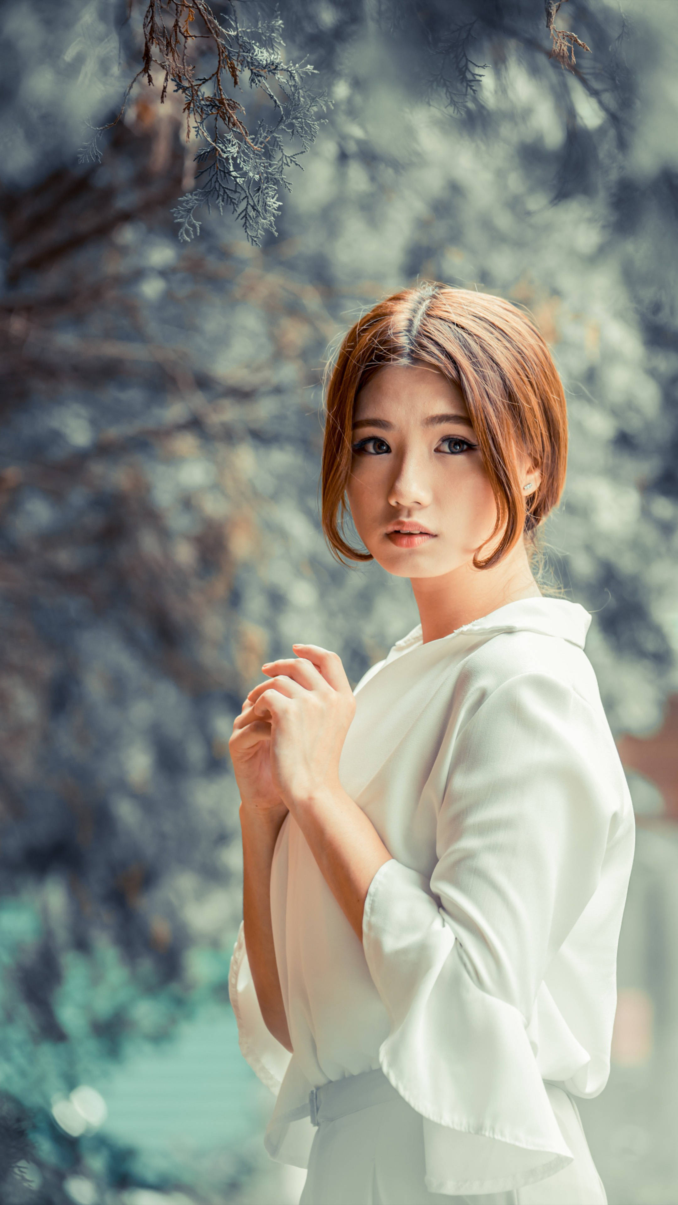 電話女孩 白裙 壁紙 — 一位身穿白色襯衫的年輕女性在柔和的自然光下優雅地站在戶外，模糊的樹葉環繞著她。