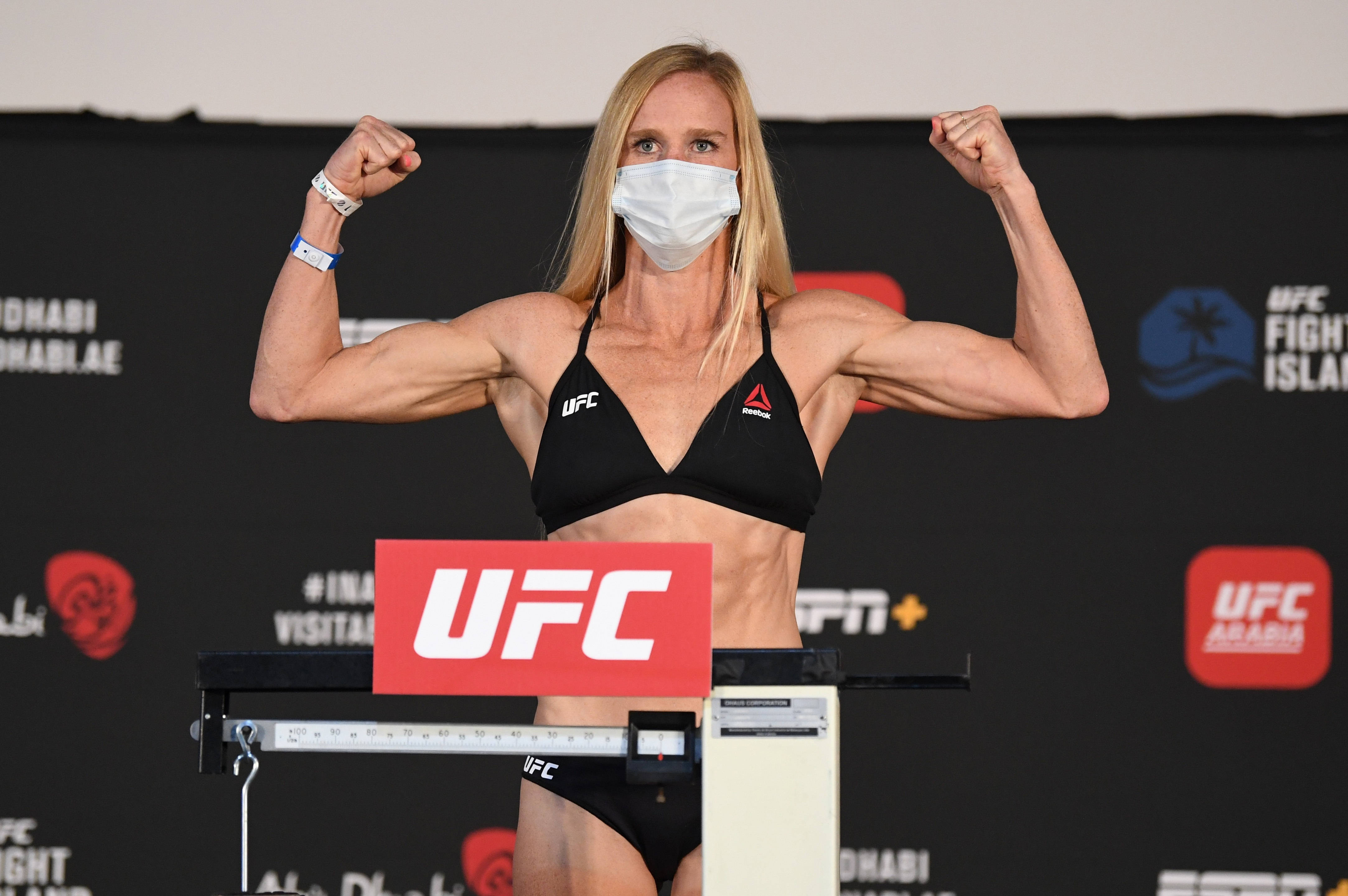 霍莉·霍姆展示肱二頭肌 壁紙 — 一位女性UFC拳手在稱重活動中戴著口罩，在體重秤上展示她肌肉發達的手臂。