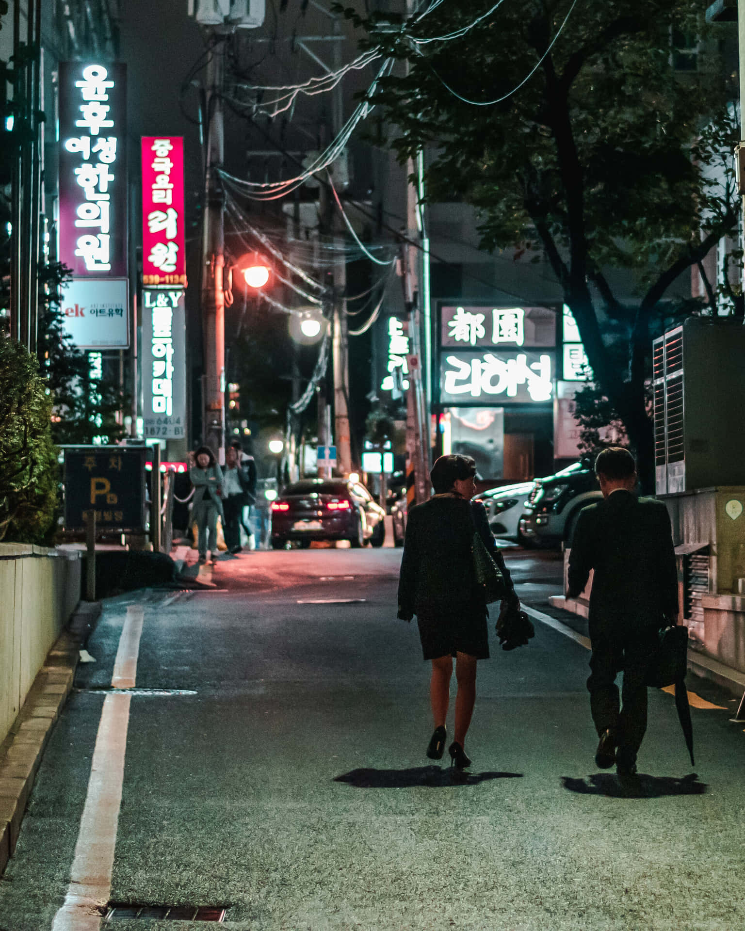 兩個人在雨夜中走在一條霓虹燈照亮的韓國城市街道上。