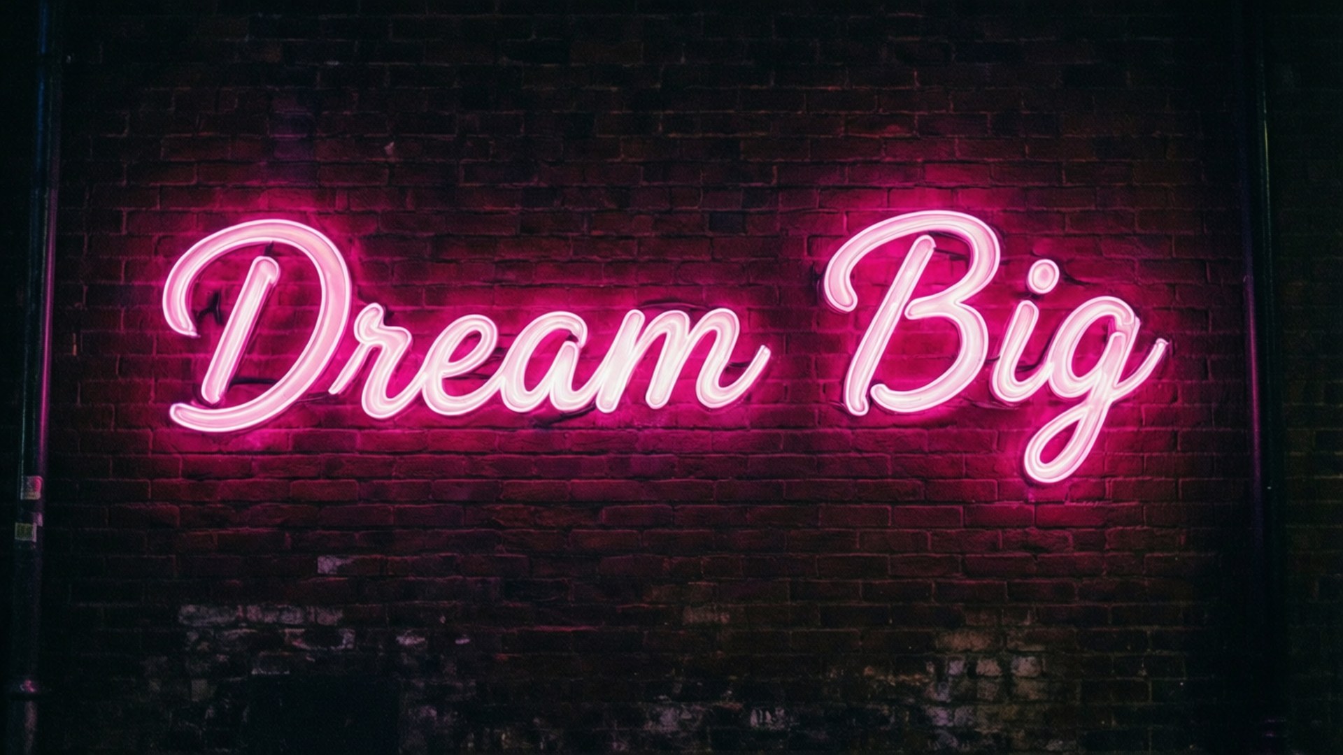 霓虹夢想大膽 壁紙 — 一個發光的粉紅色霓虹燈牌，上面寫著 Dream Big，在夜晚將鮮豔的光芒投射在深色紋理磚牆上。
