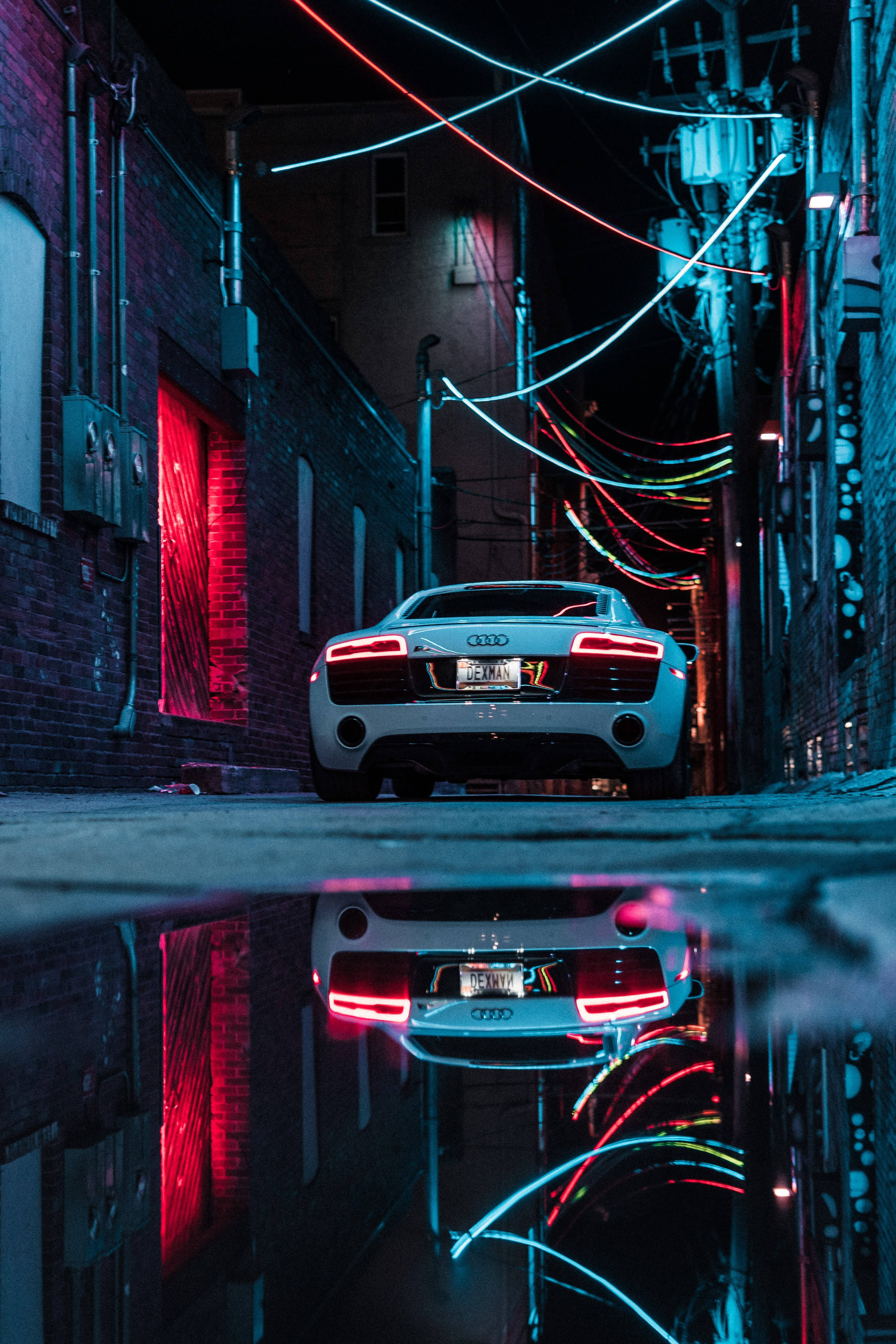 霓虹奧迪跑車 壁紙 — 夜晚，一條狹窄的城市小巷中，一輛白色跑車在霓虹燈下的水坑中倒映。