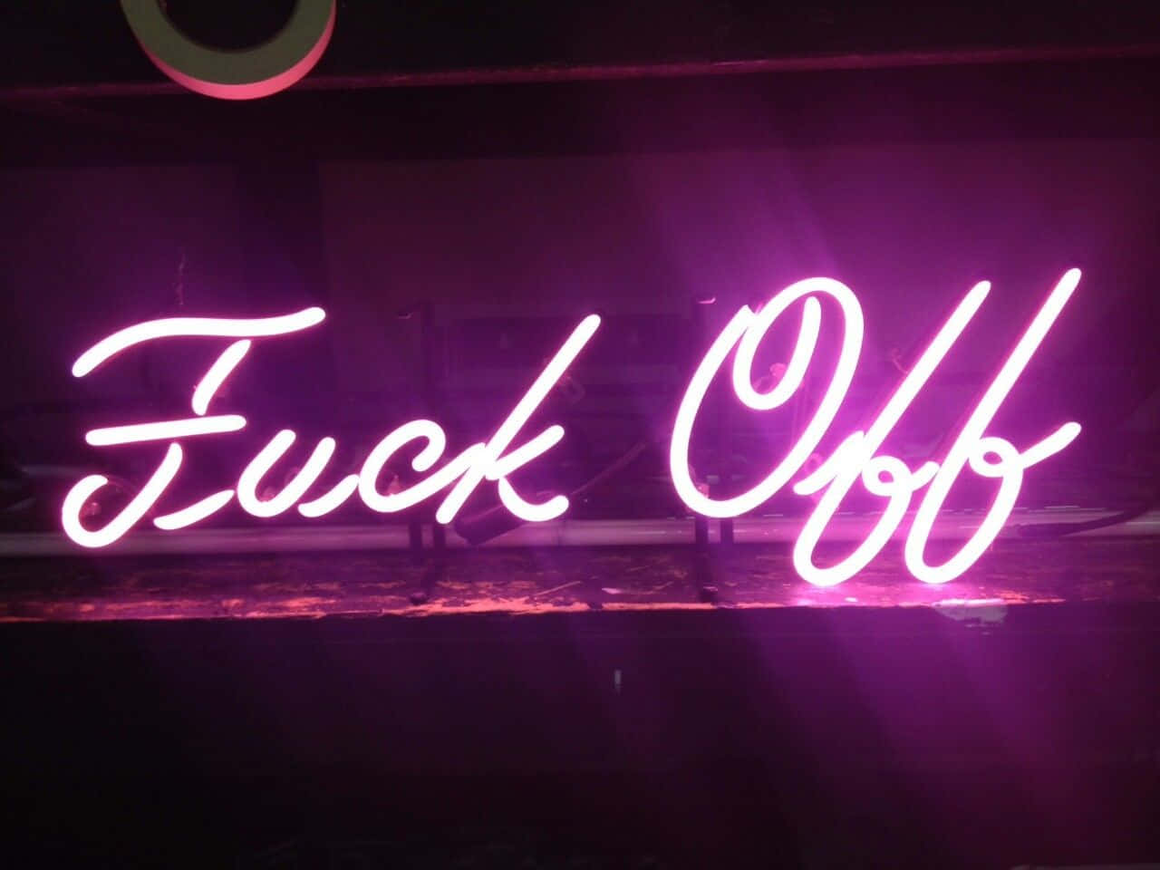 粉紅色霓虹燈牌，在黑暗背景上以草書顯示 'Fuck Off'。