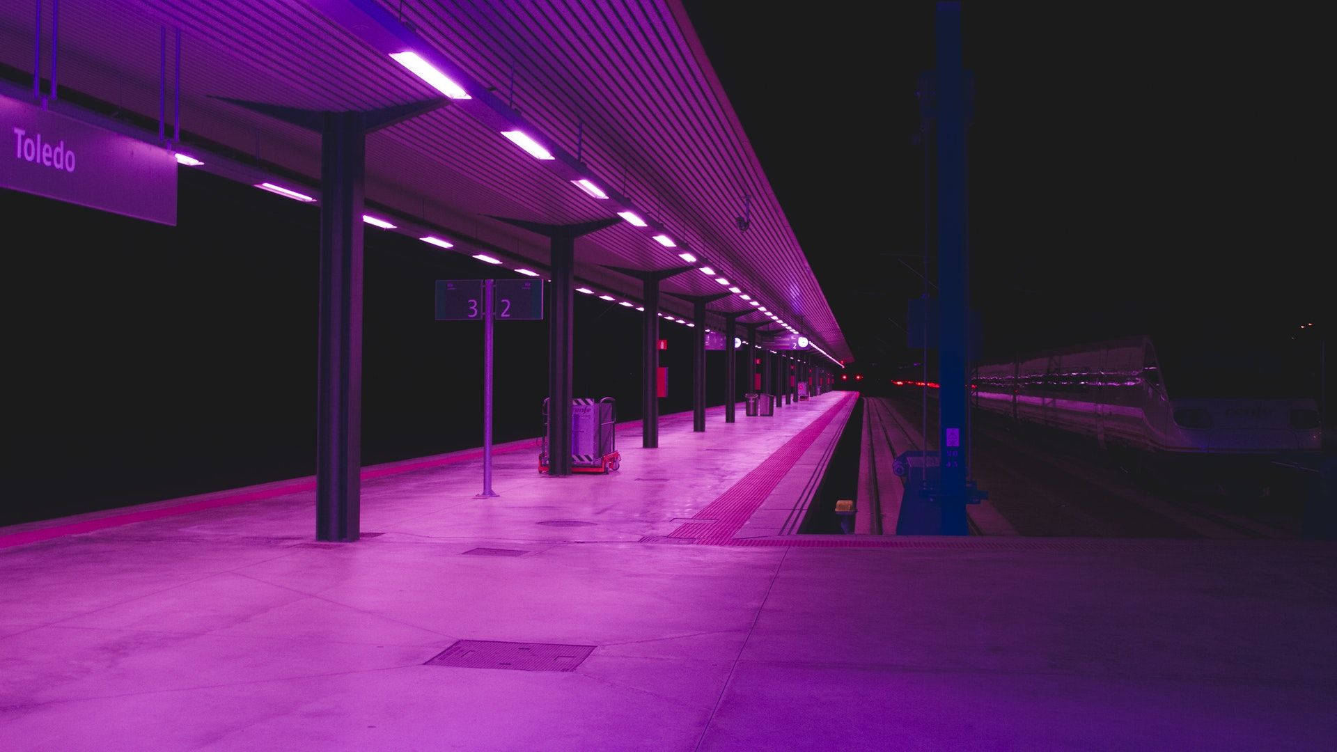 夜晚紫色燈光下的火車站月台，有經過的火車和行李車。
