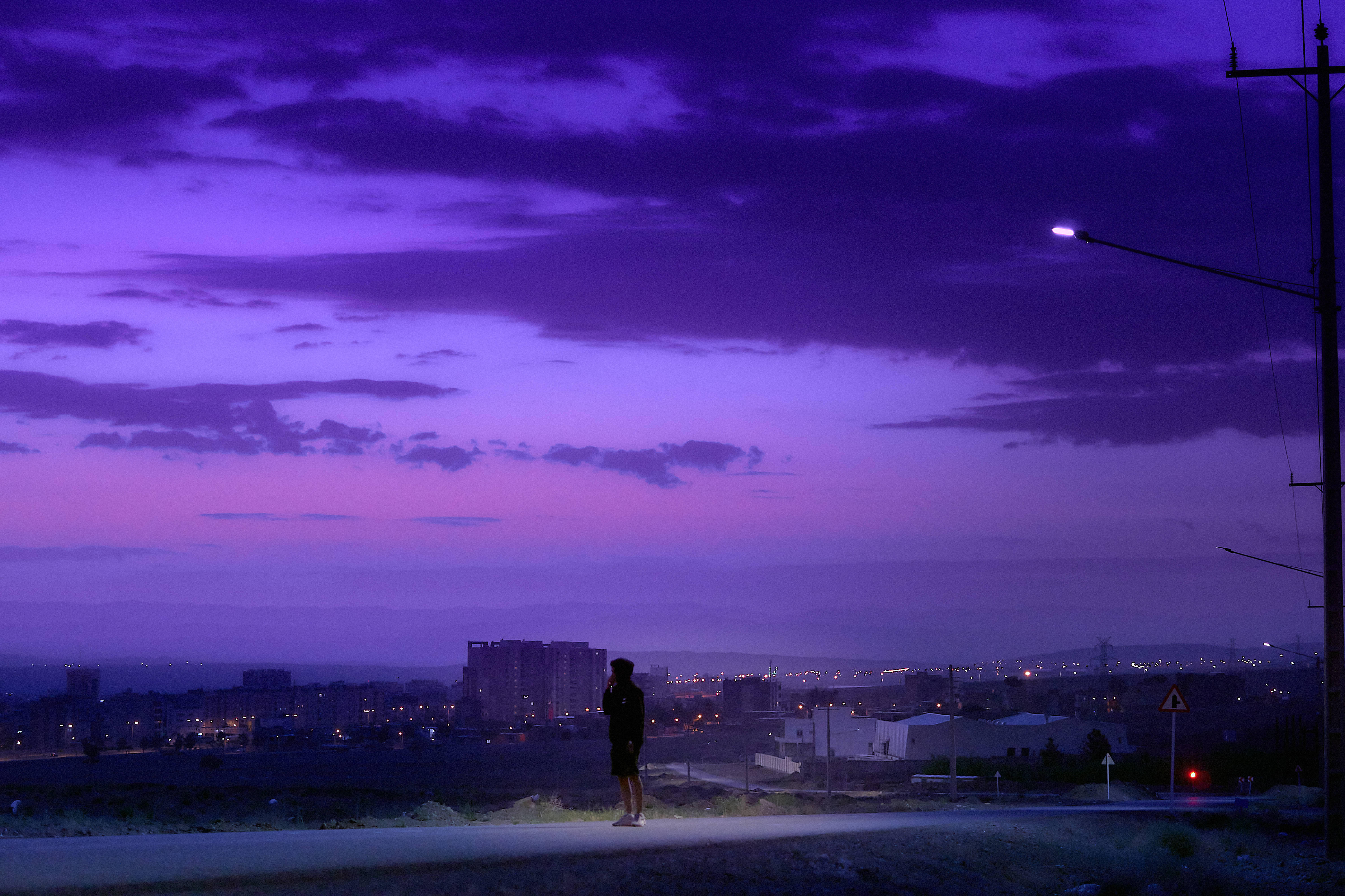 霓虹紫調夜空 壁紙 — 暮色中，一個孤獨的身影站在路邊，紫色和粉紅色的雲彩在遠處的城市景觀上方發光。