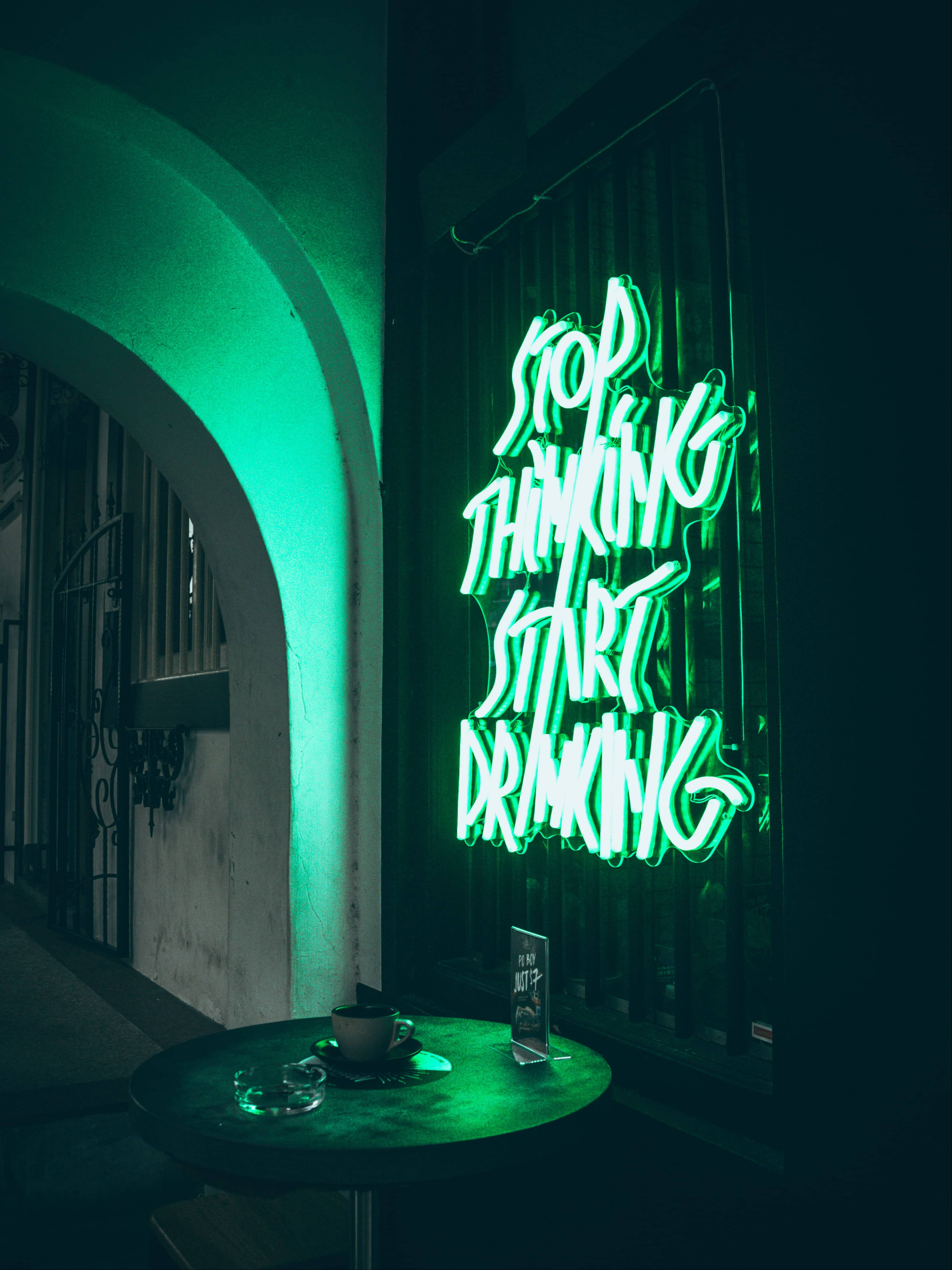 霓虹綠LED語錄招牌 壁紙 — 一個發光的綠色霓虹燈牌寫著 stop thinking start drinking，照亮了一個光線昏暗的酒吧角落，有一張小桌子放著咖啡杯和煙灰缸。