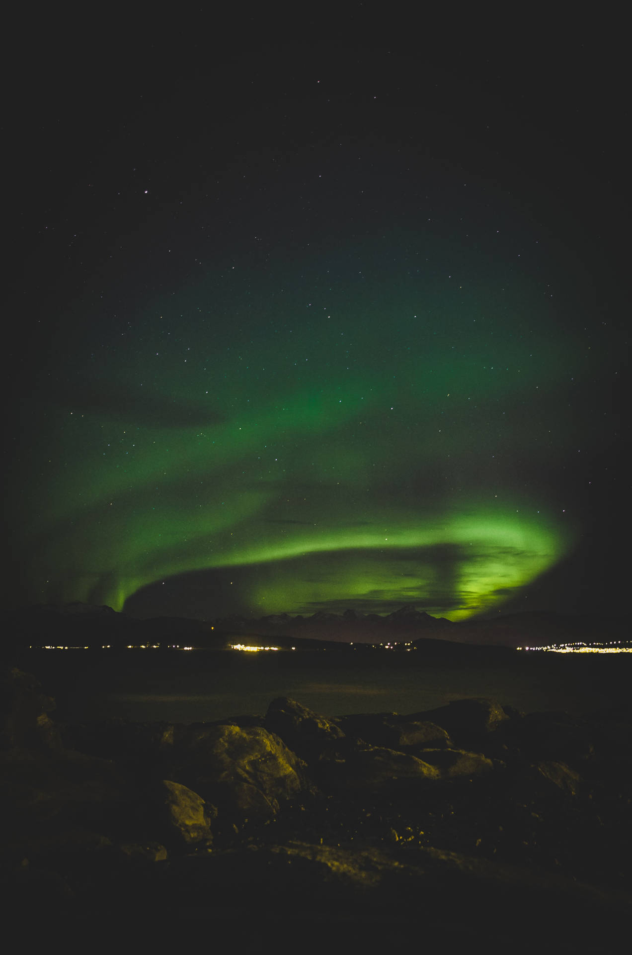 綠色北極光在黑暗的沿海景觀和遠處城市燈光上方閃爍。