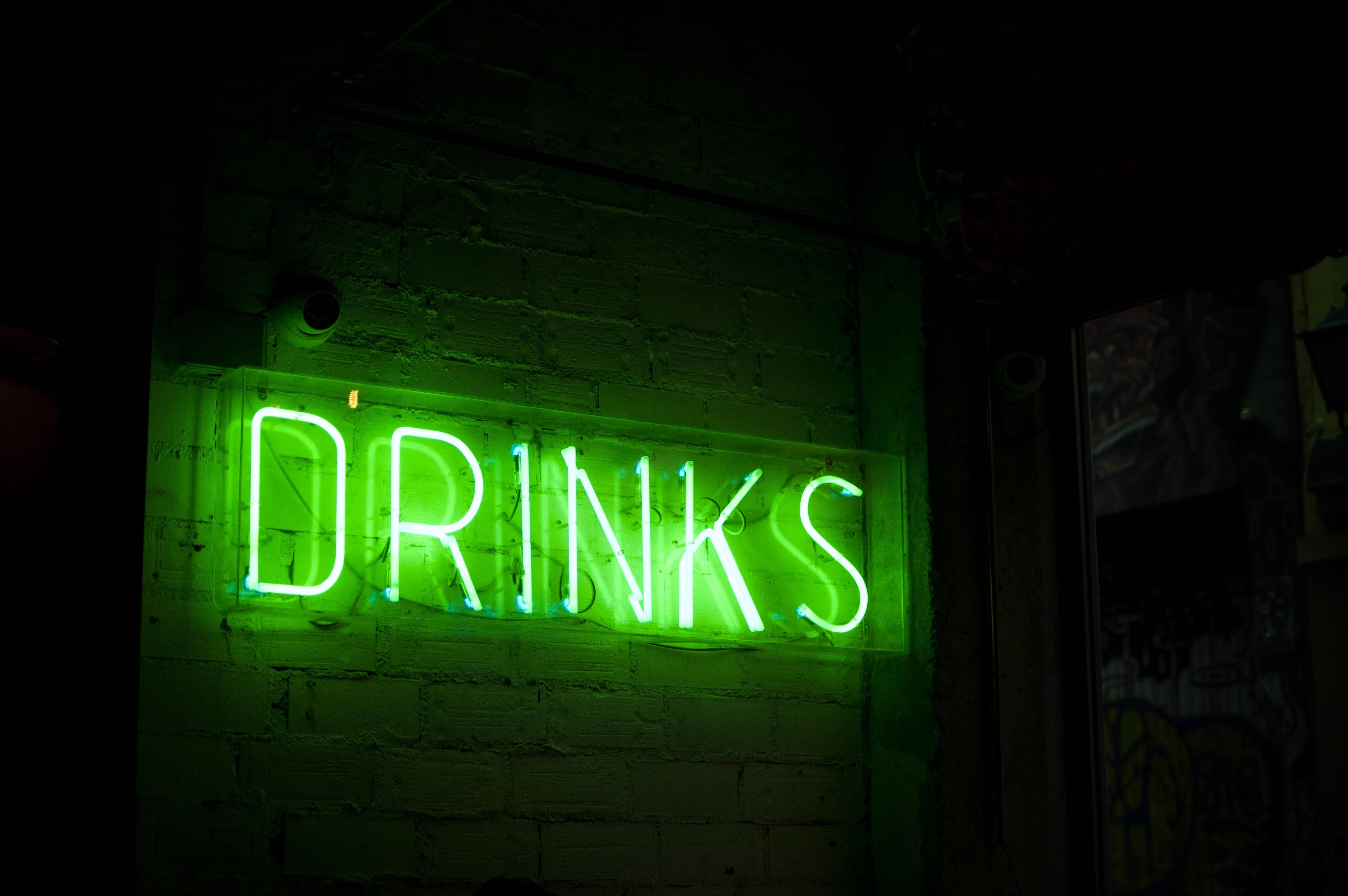 霓虹綠飲品招牌 壁紙 — 一個發光的綠色霓虹燈牌，拼寫著DRINKS，在昏暗的城市小巷中，將電光般的光芒投射在粗糙的磚牆上。