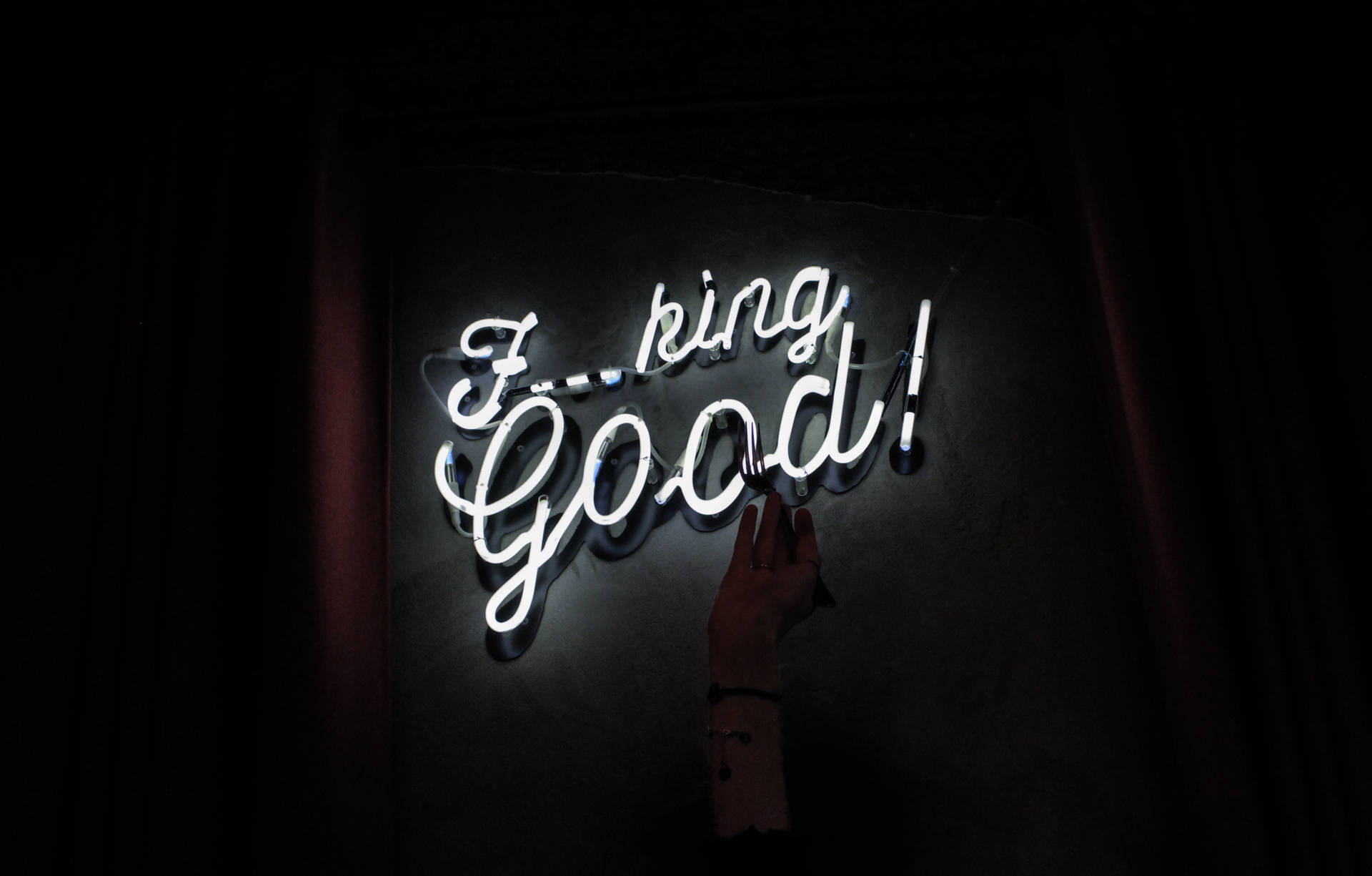 一隻手輕輕觸摸一個明亮的白色霓虹燈牌，上面寫著「F*ing Good!」，在昏暗的房間裡。