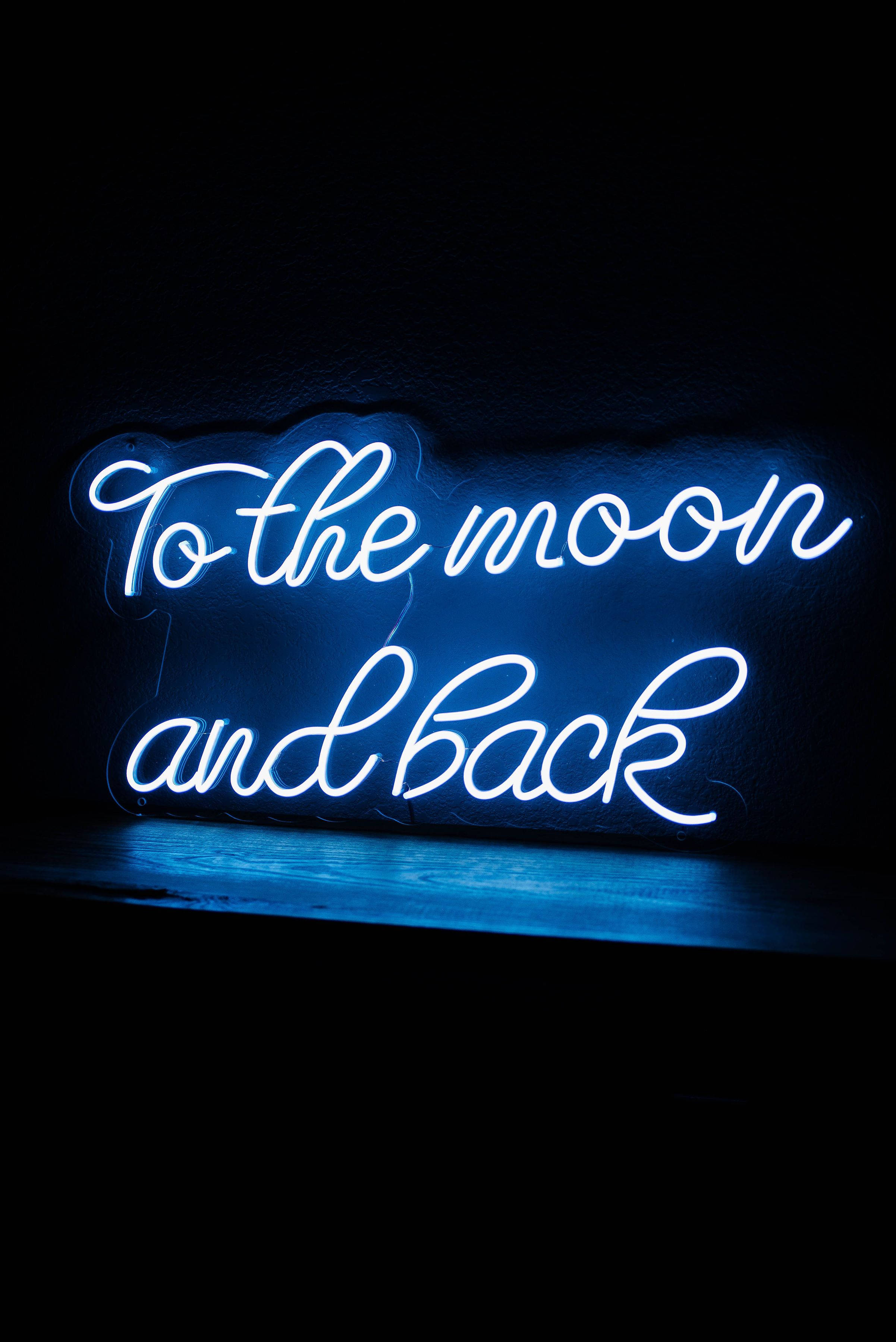 霓虹藍調情語標語 壁紙 — 一個發光的藍色霓虹燈牌，上面寫着 'To the moon and back'，背景是深色紋理牆壁，下方有反射表面。