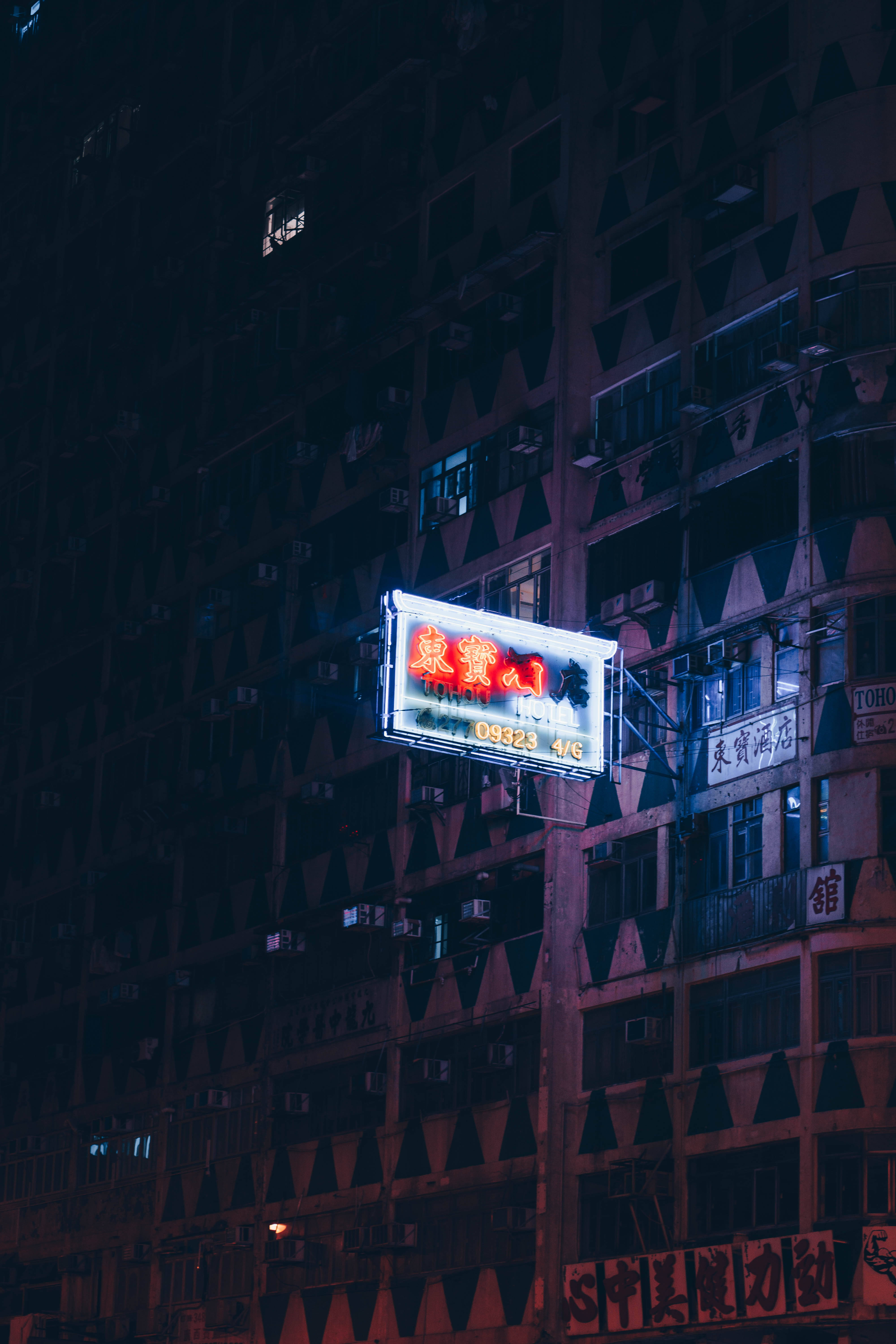 霓虹酒店招牌 iPhone 壁紙 — 一個發光的霓虹燈牌在夜晚照亮了密集的城市建築外牆，將溫暖的光線投射在黑暗的幾何圖案上。