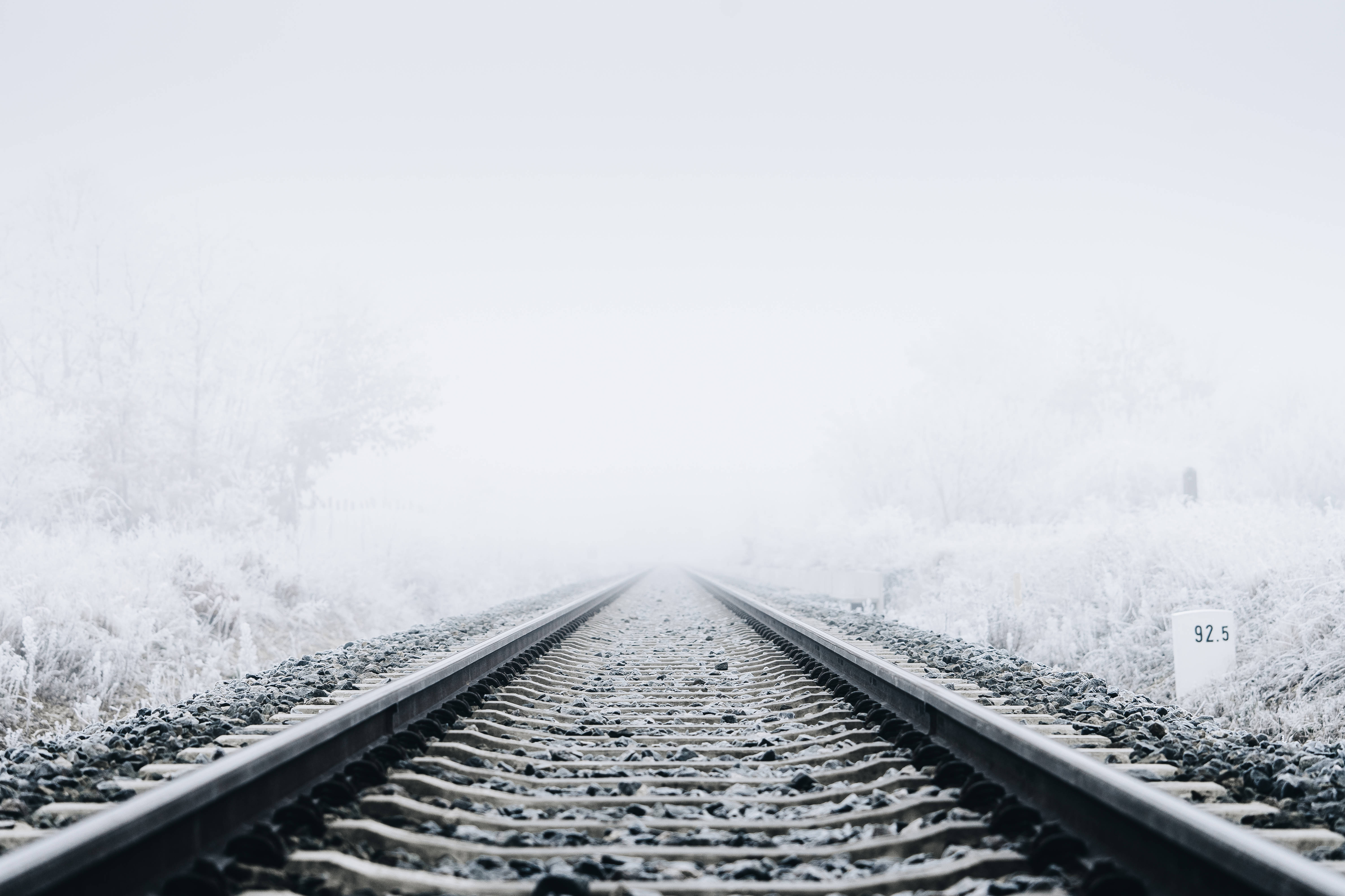 霧中鐵路軌道冬季桌面 壁紙 — 被雪覆蓋的鐵路軌道延伸進一個霧濛濛的白色冬季景觀，兩旁是結霜的樹木。