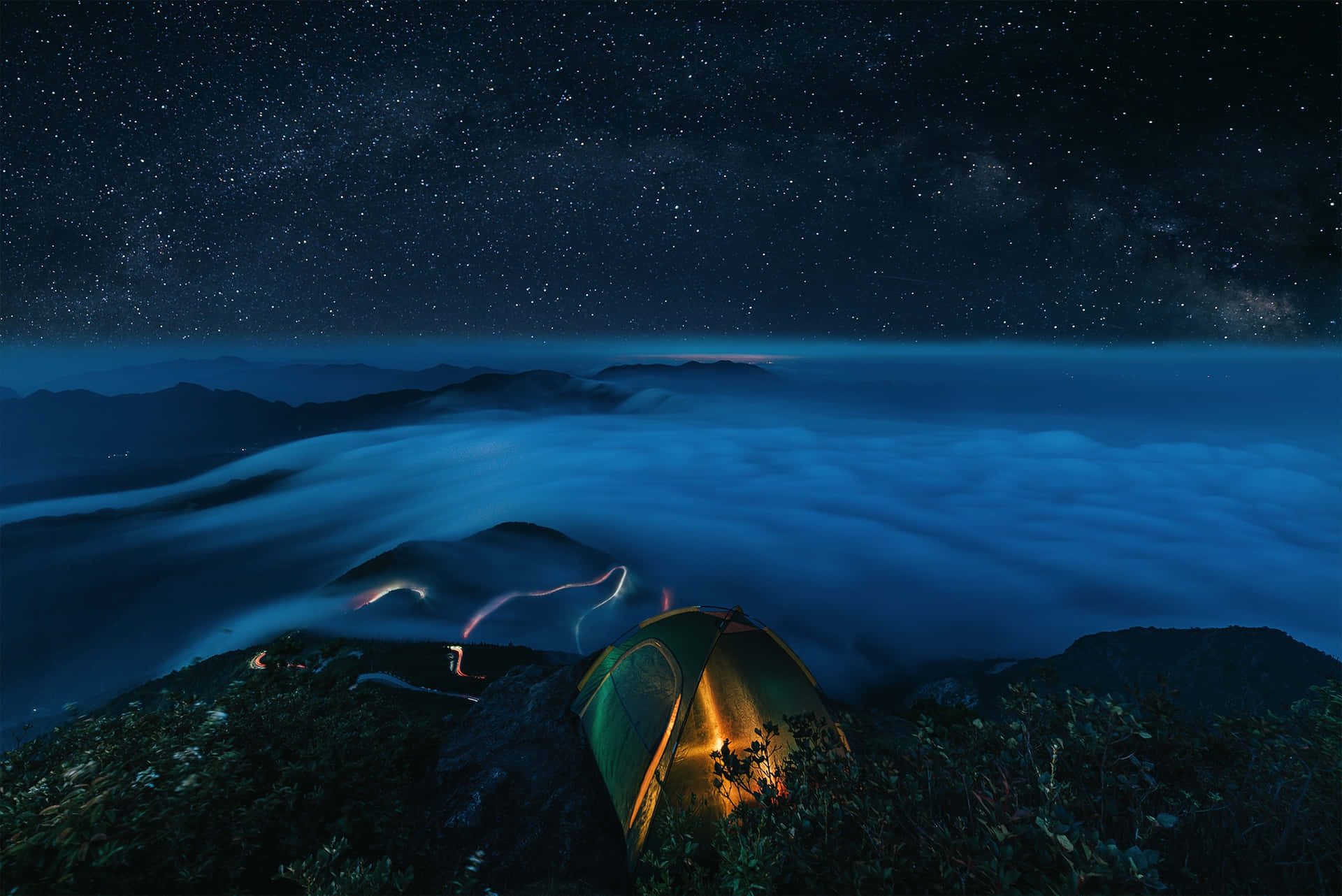 夜間山上的亮燈帳篷，下方有星星和雲。
