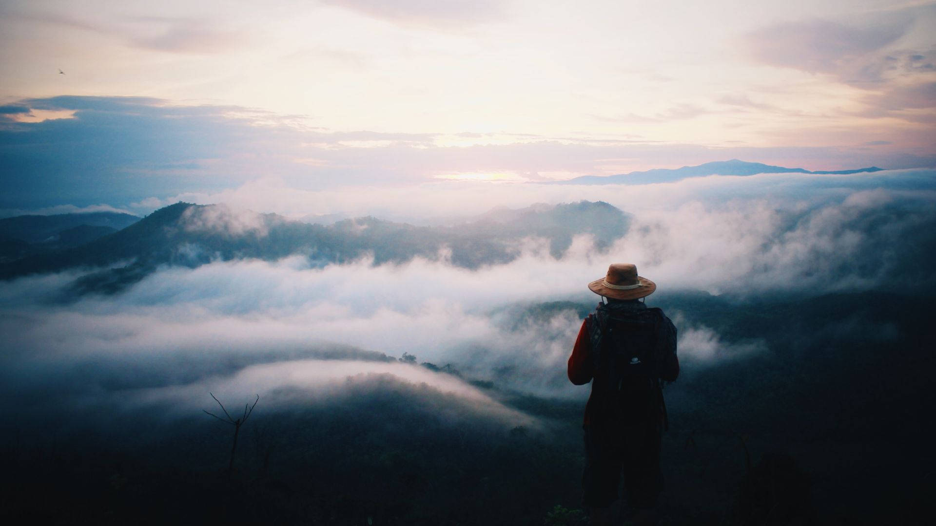 霧山中的人類旅程 壁紙 — 一位戴草帽的孤獨遠足者站在山脊上，俯瞰黎明時分的雲海。