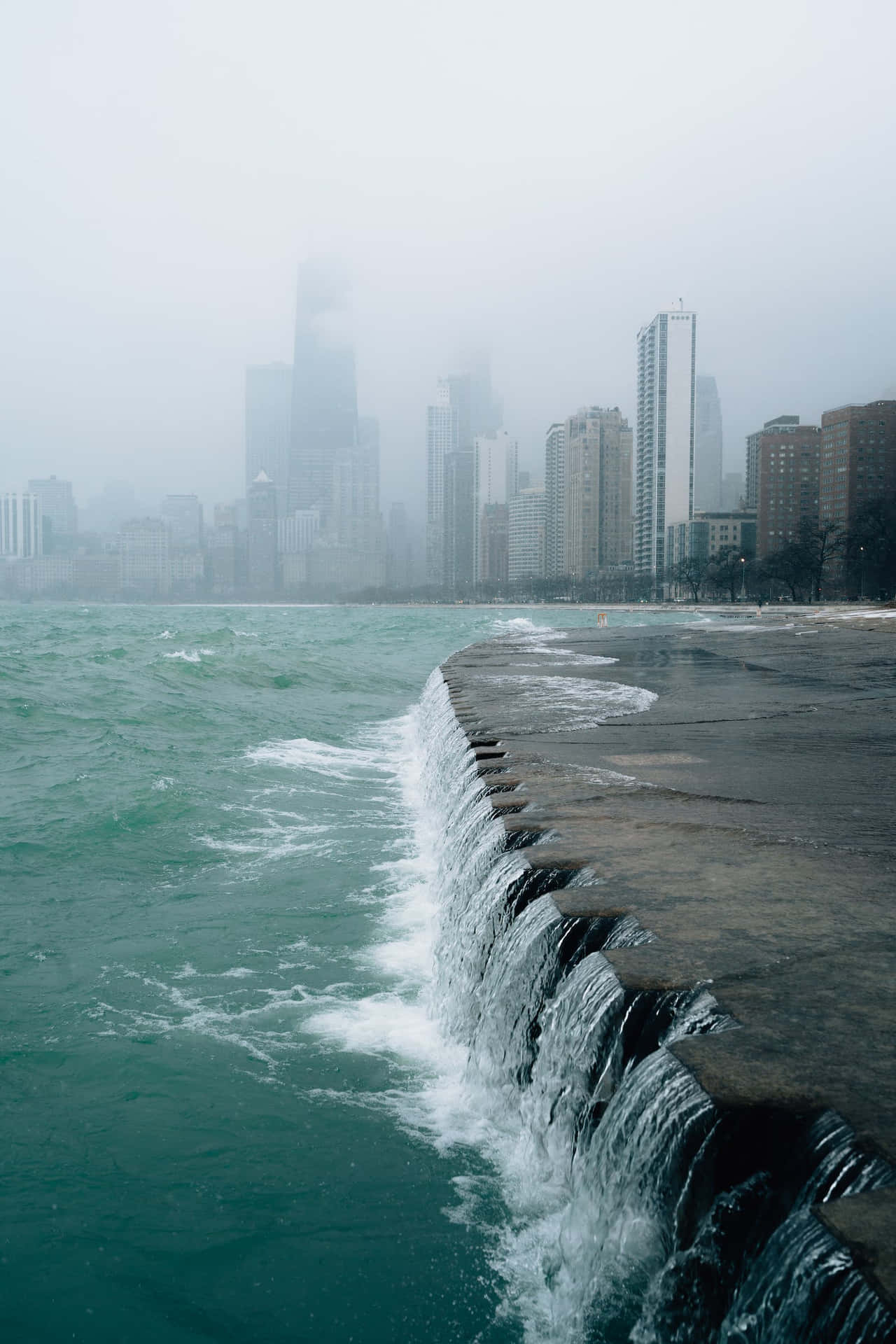 海浪拍打石堤，背景是霧氣瀰漫的城市天際線。