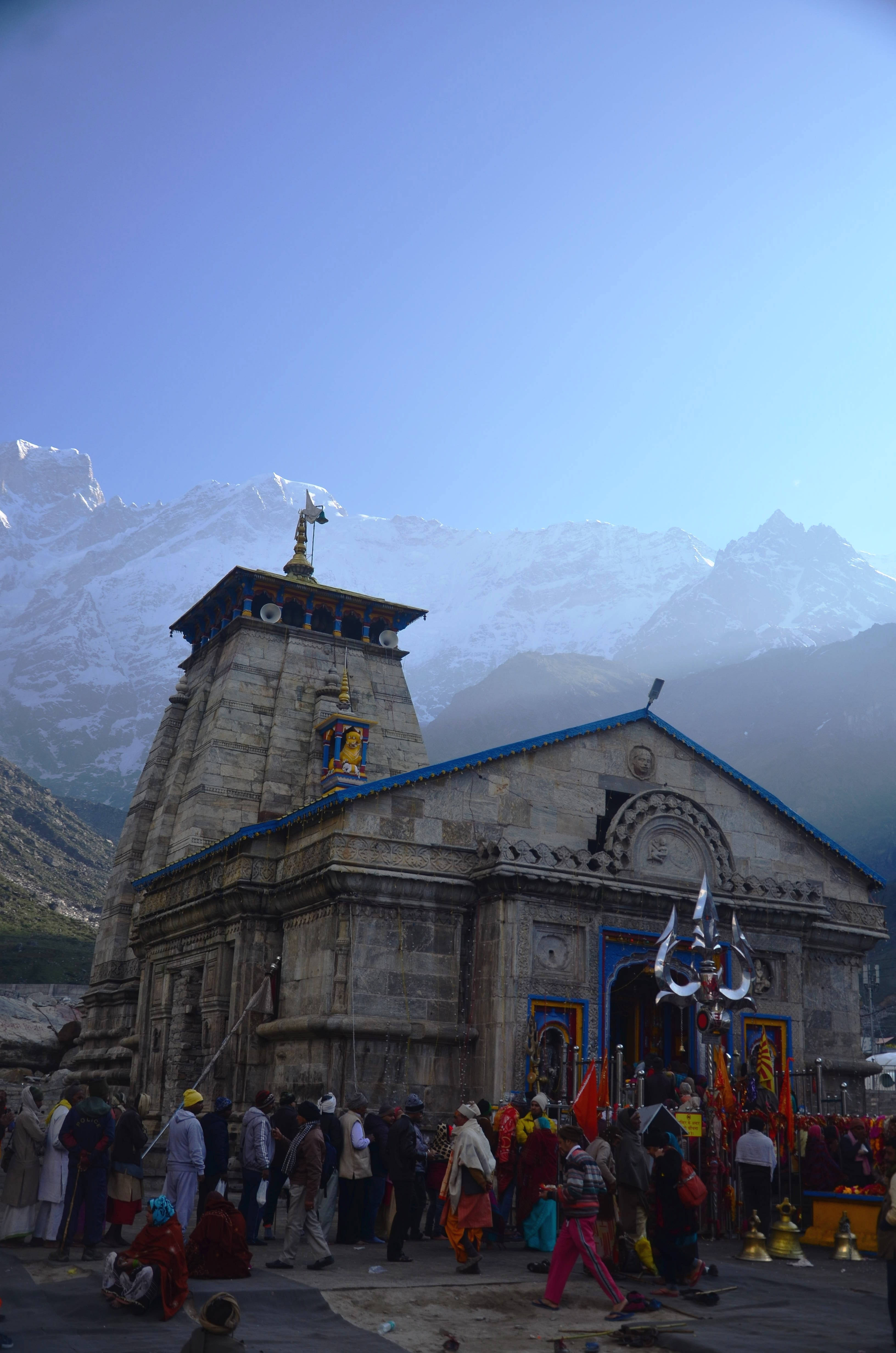 霧氣克德納特寺廟 4K 壁紙 — 一座藍色屋頂的石廟矗立在積雪的山峰前，朝聖者在晴朗的藍天下聚集在入口處。
