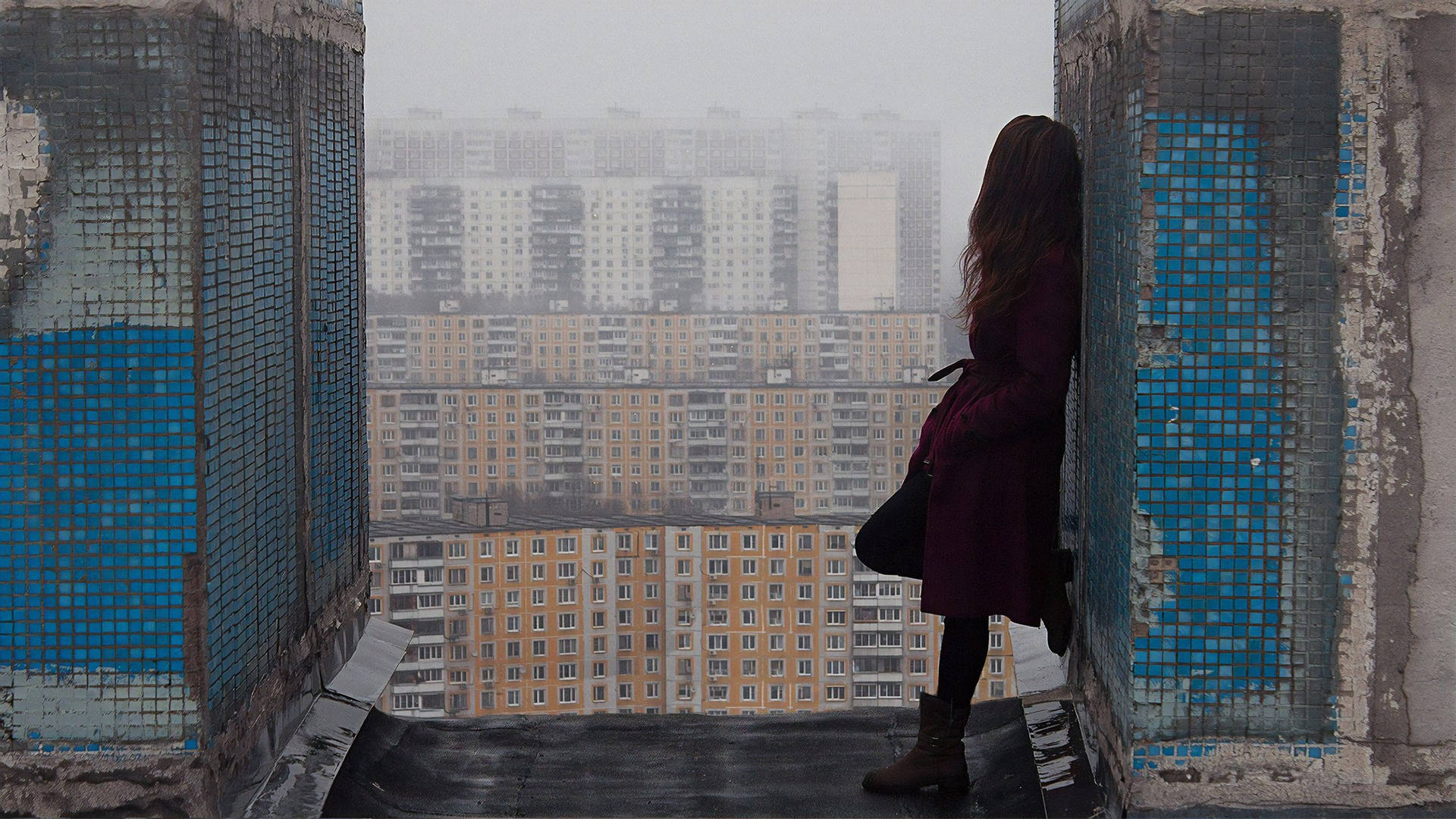 霧氣瀰漫的工業城市風景 壁紙 — 一位身穿栗色外套的孤獨女子站在風化的屋頂邊緣，由藍色馬賽克瓷磚框住，俯瞰著朦朧的城市天際線。