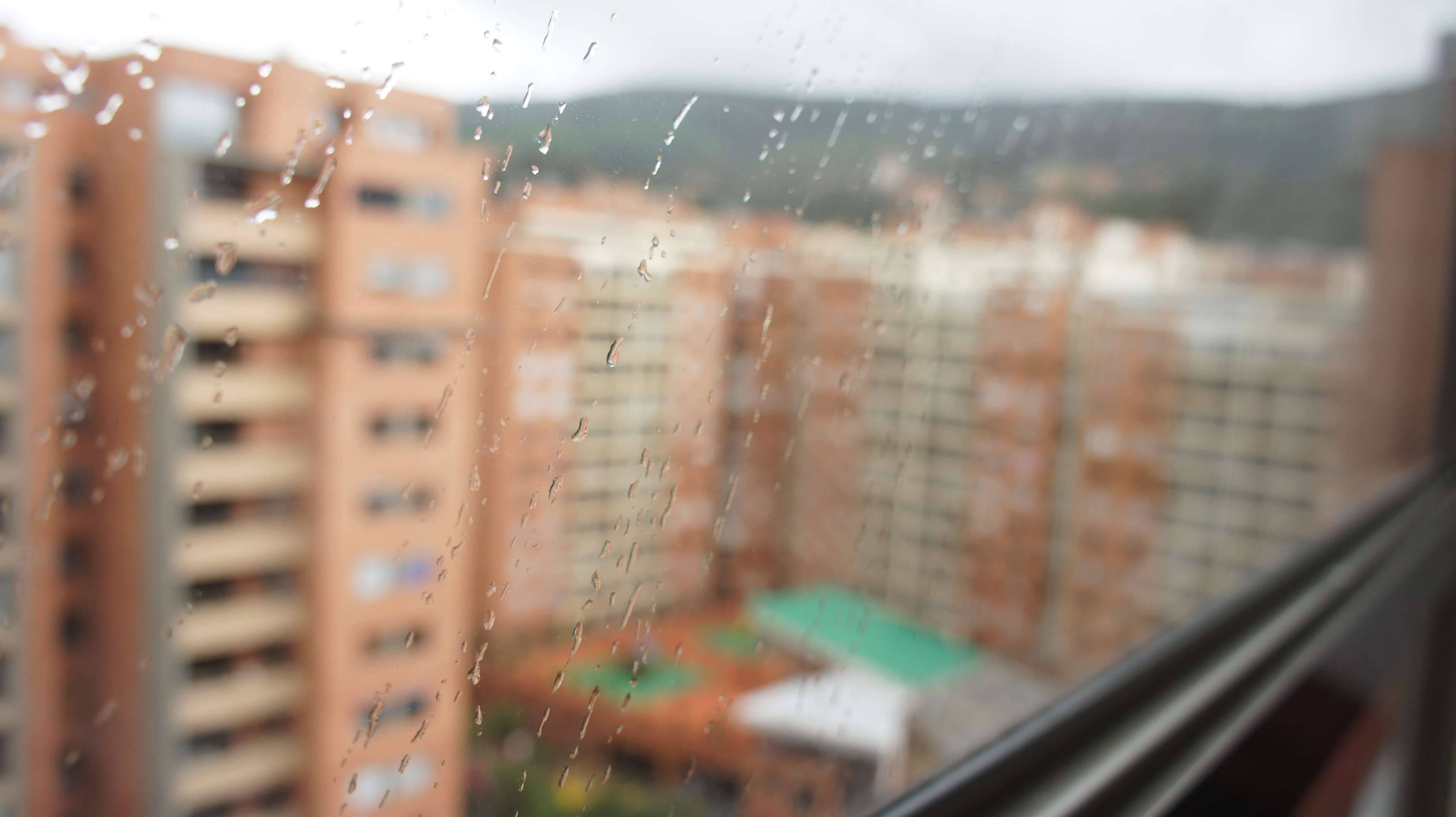 霧氣瀰漫的波哥大 壁紙 — 雨滴沿著窗玻璃流下，模糊了陰天天空下橙色公寓樓和遠處綠色山丘的景色。