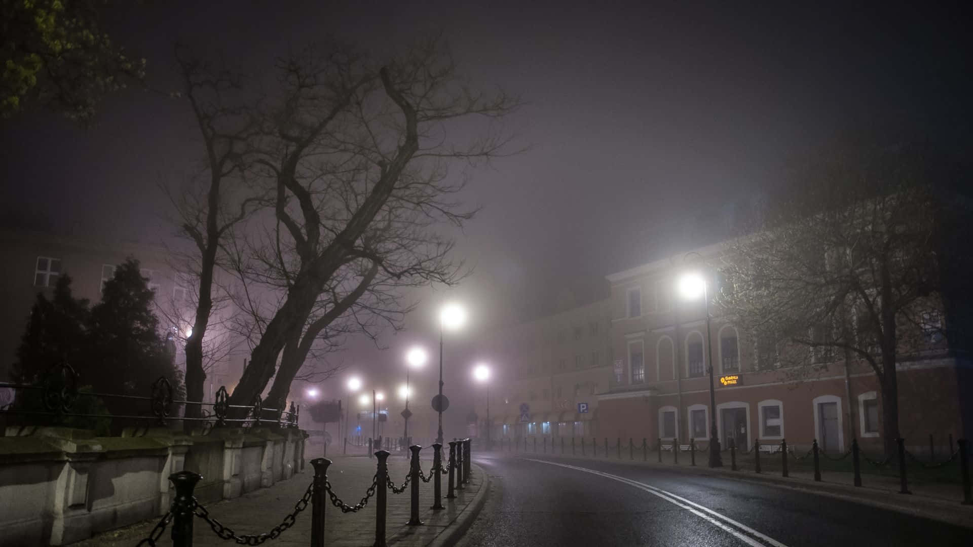 霧氣籠罩的夜間街景 壁紙 — 一條霧氣瀰漫的夜晚街道，被發光的路燈照亮，柔和的光線灑在光禿禿的樹木和磚砌建築上。