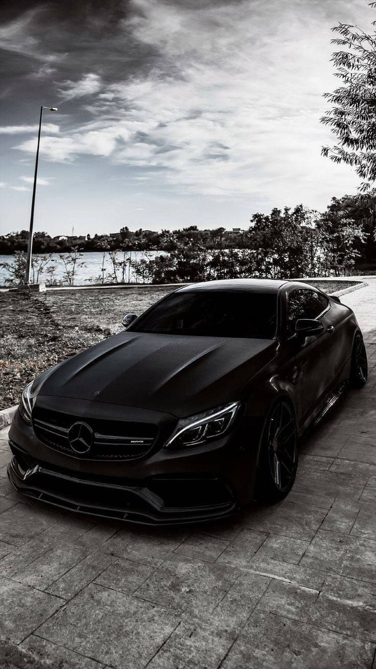 霧面黑色梅賽德斯-奔馳AMG 壁紙 — 一架時尚的啞光黑色Mercedes-AMG跑車停泊在平靜湖泊旁的石徑上，天空多雲。