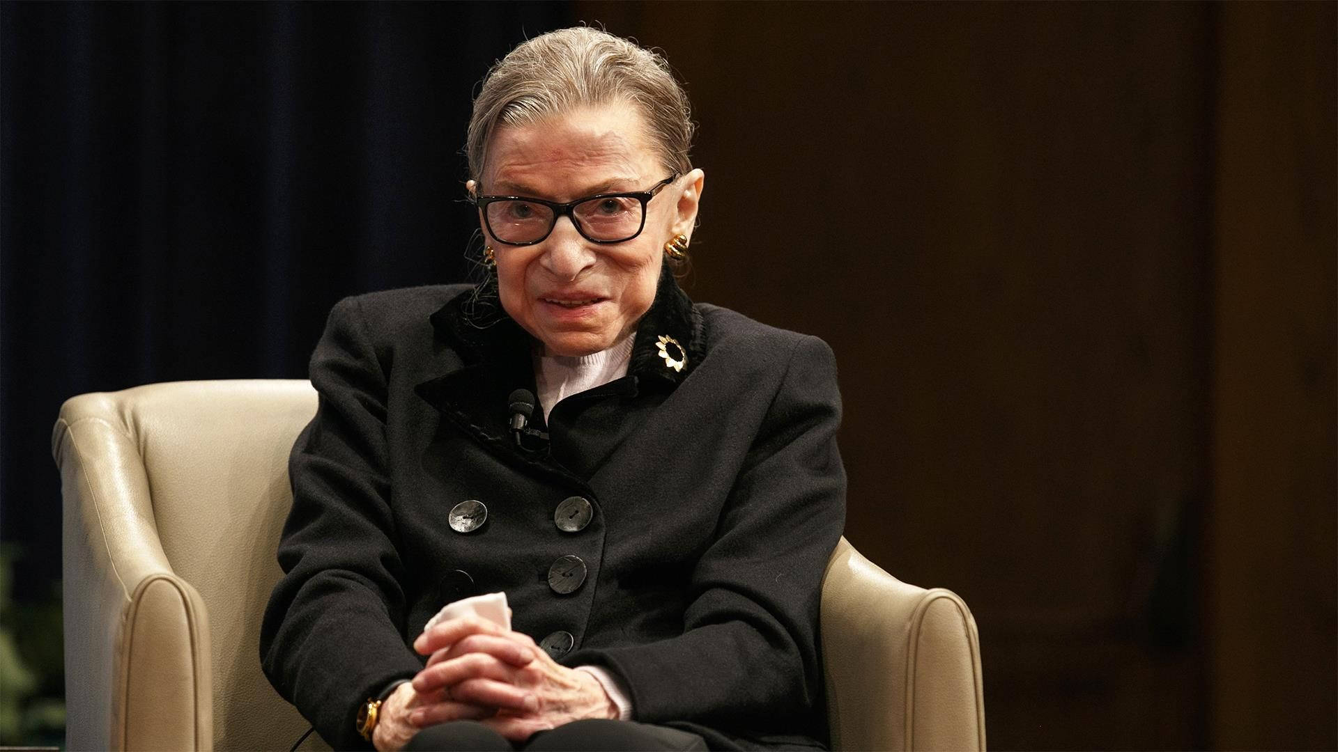 露絲·巴德·金斯伯格坐在沙發上 壁紙 — Ruth Bader Ginsburg 坐在米色扶手椅上，穿著黑色外套和眼鏡，表情嚴肅。