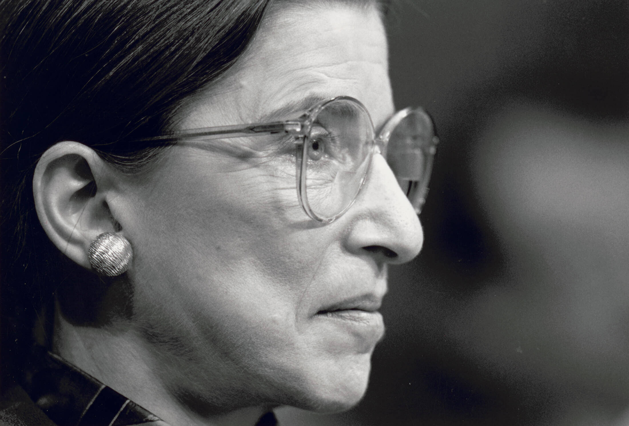 露絲·巴德·金斯伯格灰階照片 壁紙 — Ruth Bader Ginsburg 的側面黑白特寫肖像，戴著眼鏡和獨特的耳環。