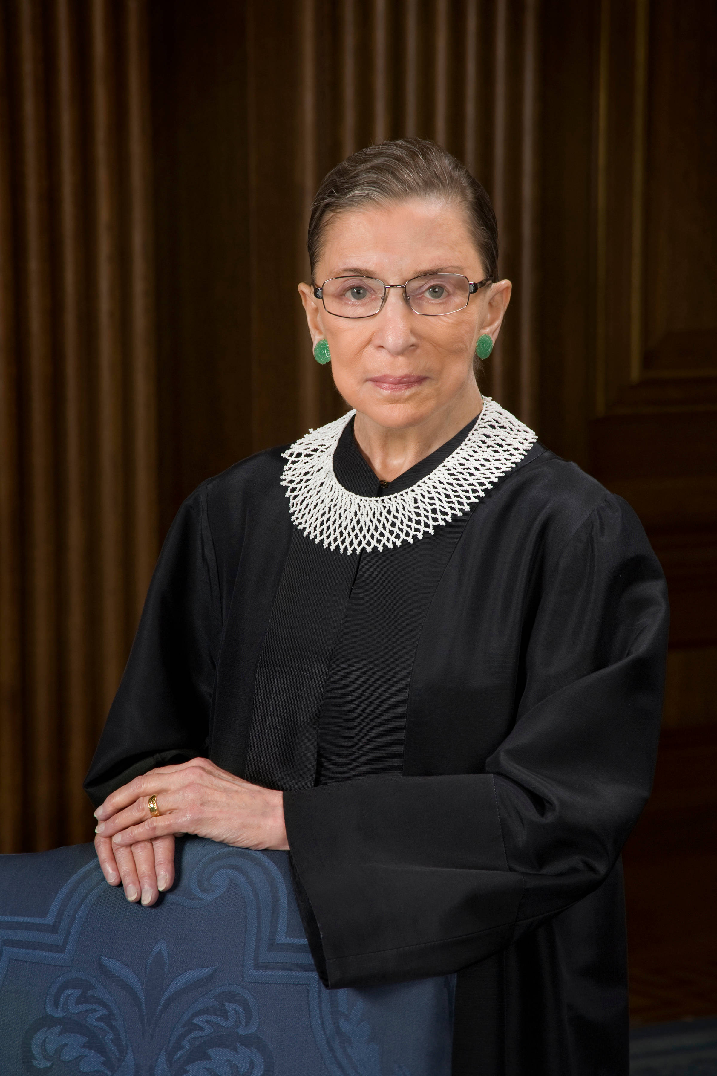 露絲·巴德·金斯伯格穿傳統黑袍 壁紙 — Ruth Bader Ginsburg 身穿黑色法官袍，搭配白色蕾絲領和綠色耳環，以木質背景拍攝的肖像。