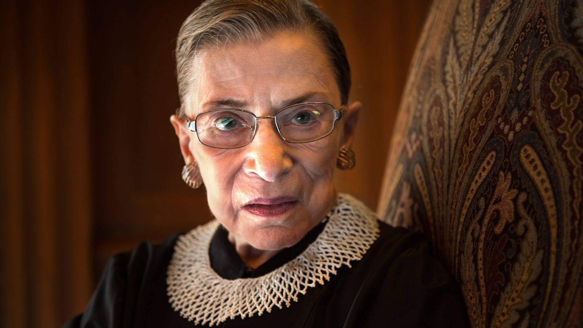 露絲·巴德·金斯伯格美國律師 壁紙 — Ruth Bader Ginsburg 的 close-up 肖像，坐在有圖案的椅子上，穿著她標誌性的黑色長袍和蕾絲領。