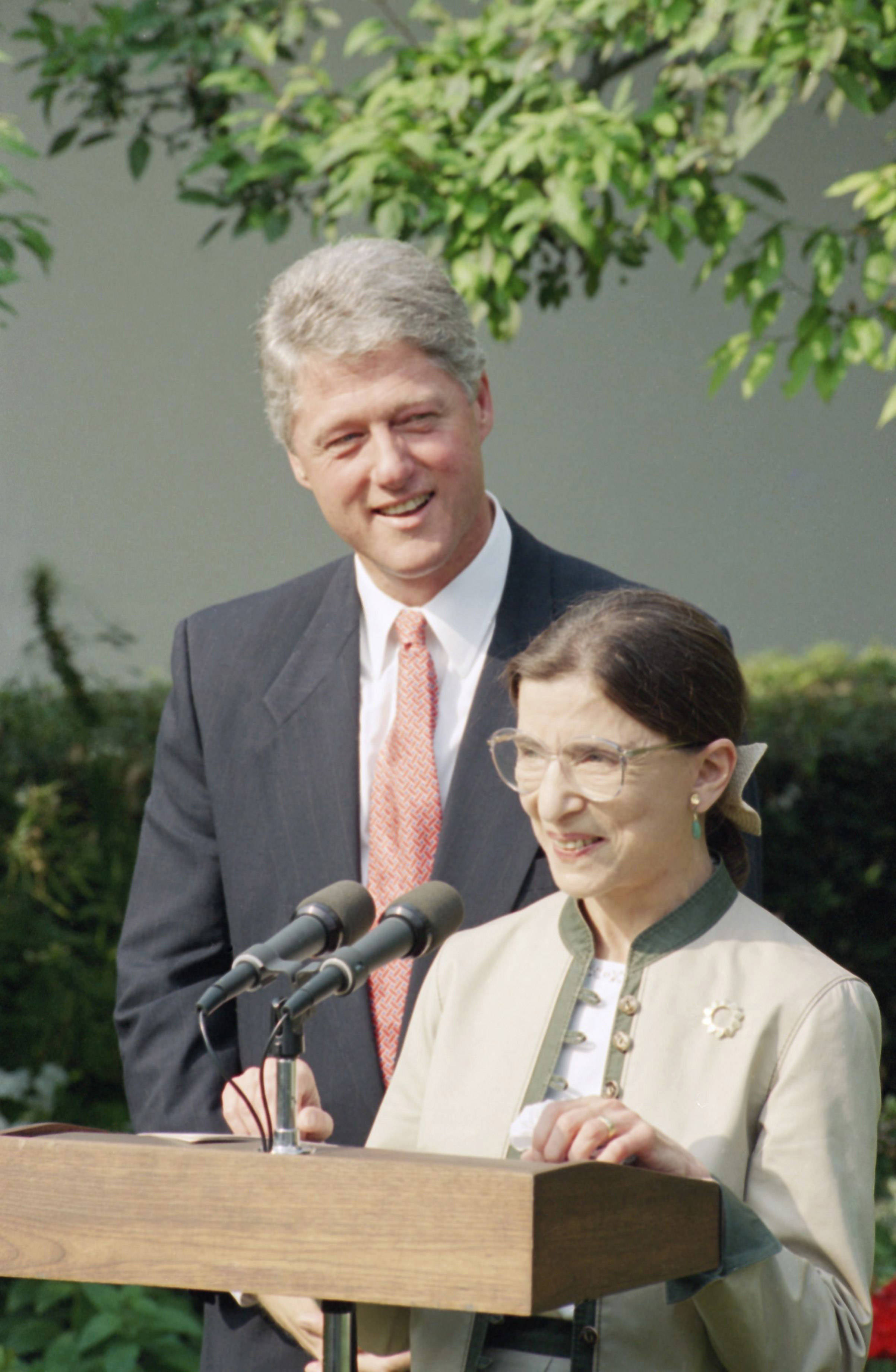 露絲·巴德·金斯伯格與比爾·克林頓 壁紙 — Ruth Bader Ginsburg 在講台上發言，比爾·克林頓在她身後綠樹成蔭下微笑。