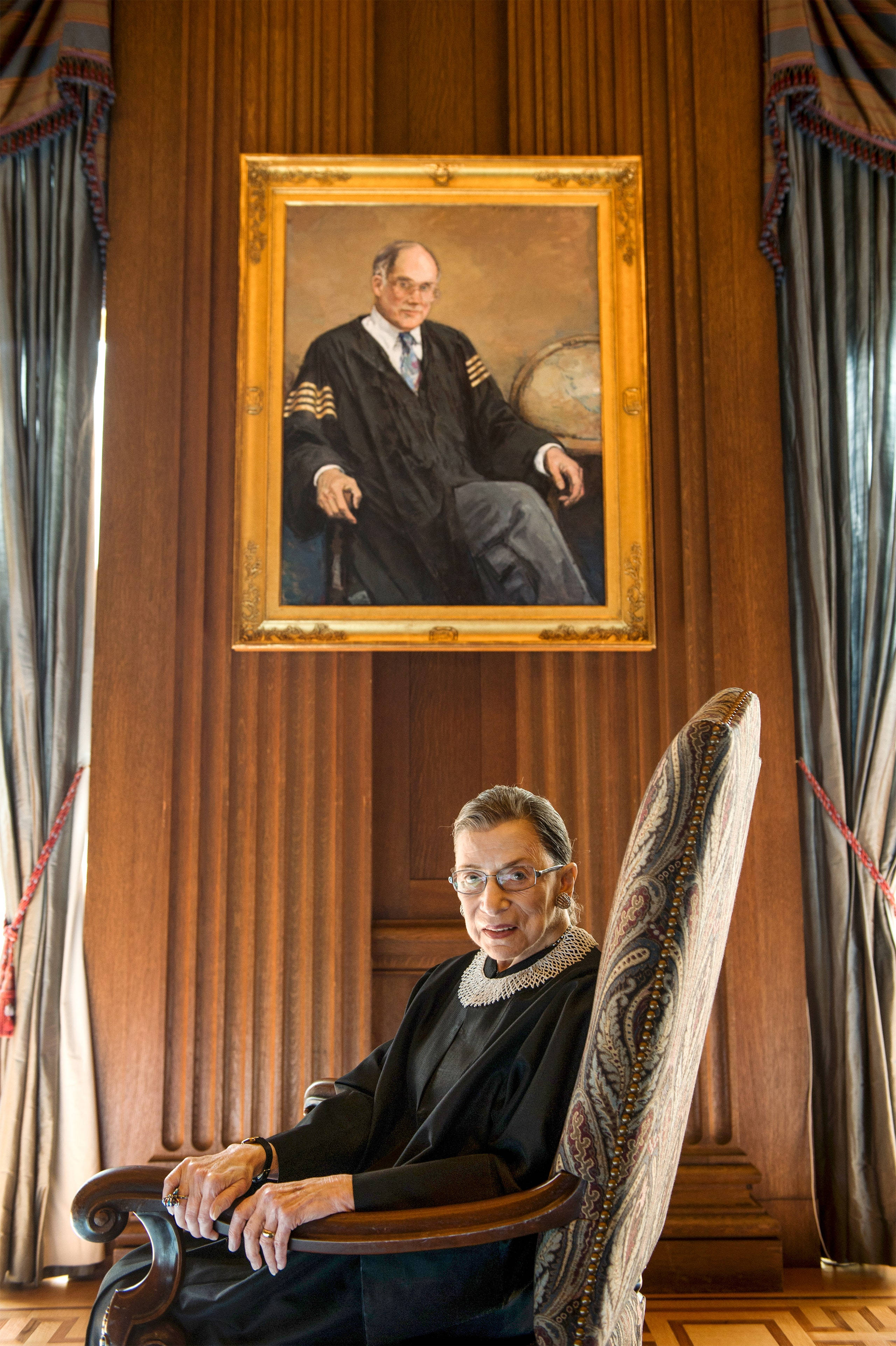 露絲·巴德·金斯伯格與畫作 壁紙 — Ruth Bader Ginsburg 坐在木椅上，下方掛著她身穿司法袍的肖像，背景是華麗的木鑲板和垂簾。