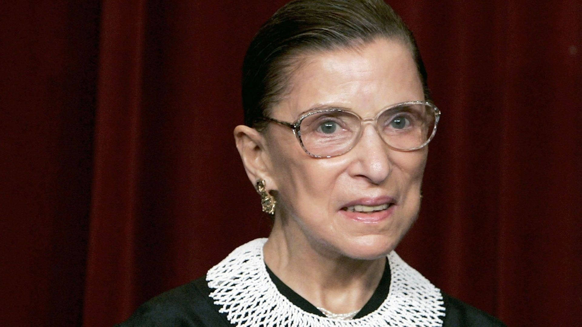 露絲·巴德·金斯伯格銀色眼鏡 壁紙 — Ruth Bader Ginsburg 身穿法官袍，以深紅色窗簾為背景的肖像。