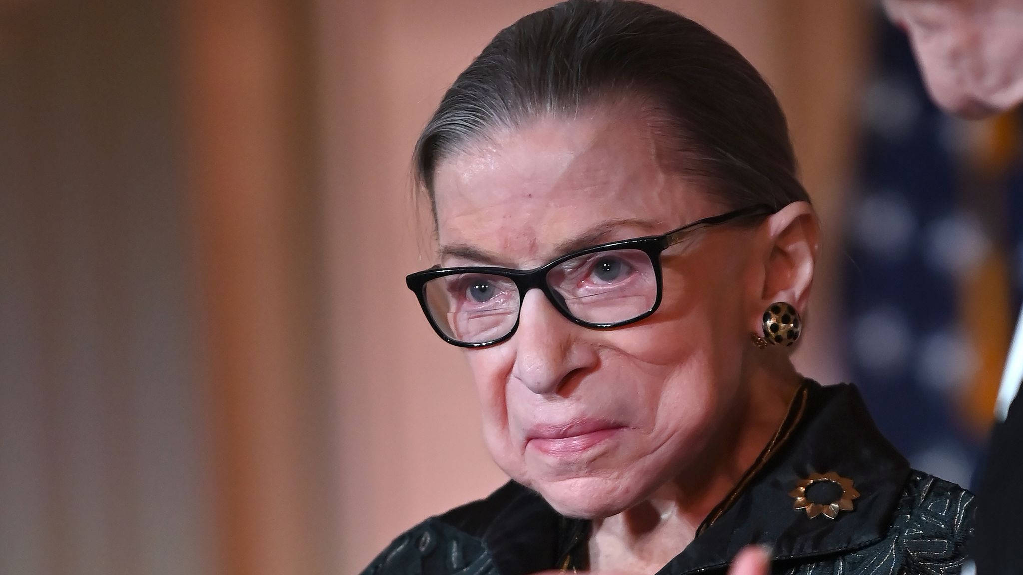 露絲·巴德·金斯伯格黑色框眼鏡 壁紙 — Ruth Bader Ginsburg 嘅特寫肖像，戴住黑色框眼鏡同深色圖案外套，上面有個太陽光線胸針。