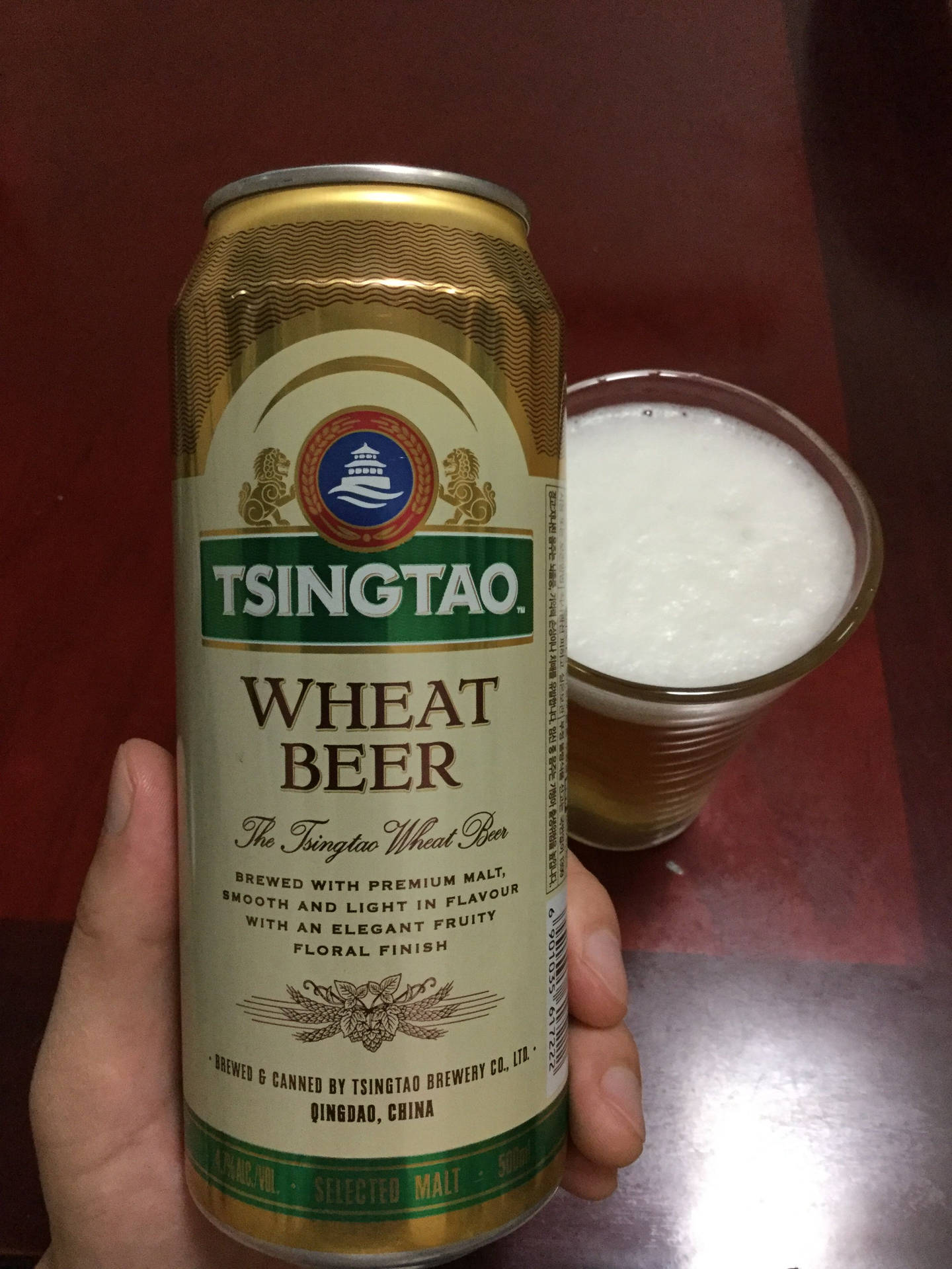 手在紅色桌子上拿著青島小麥啤酒罐，旁邊放著一杯泡沫啤酒。