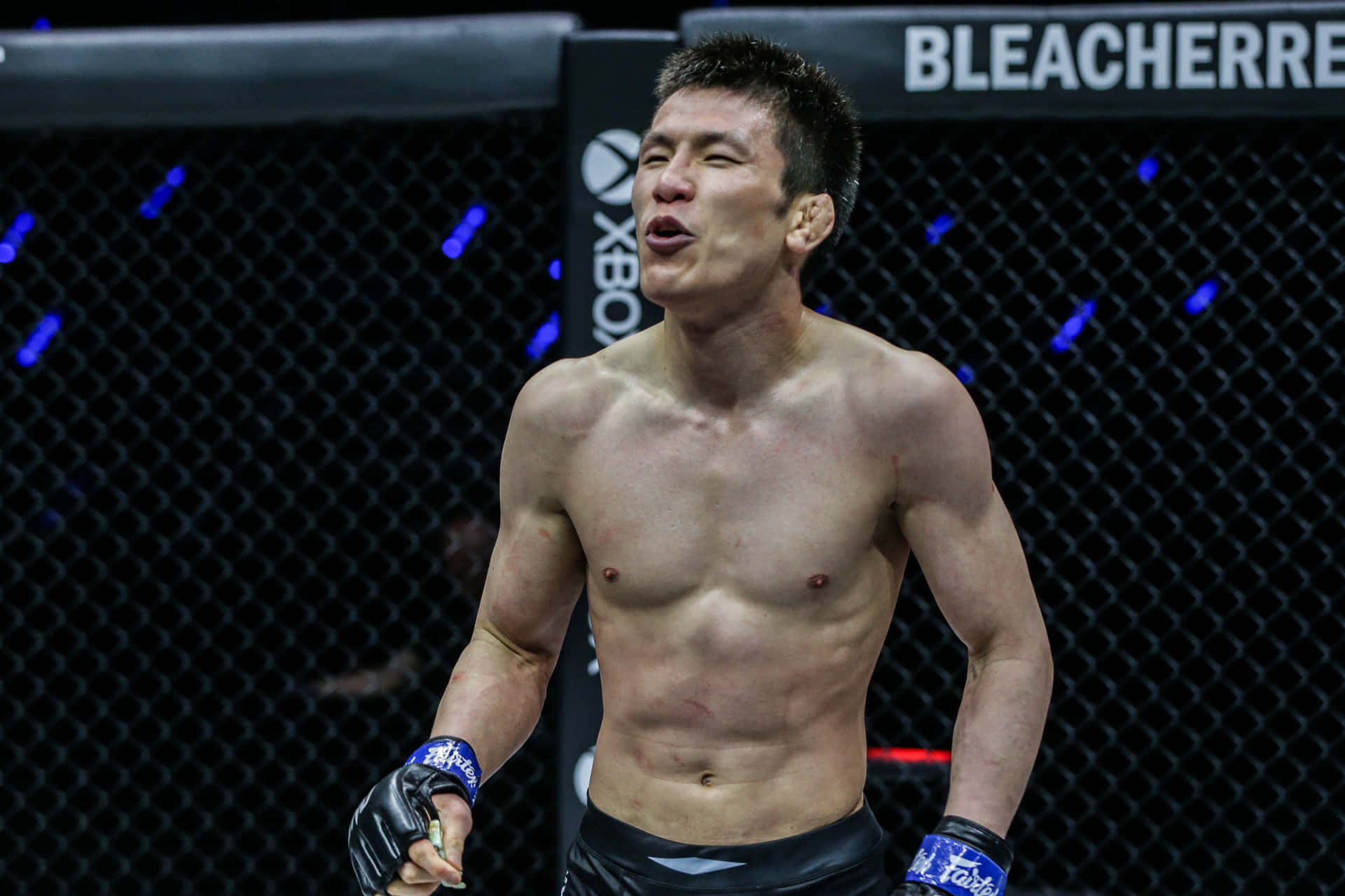 青木真也興奮加入ONE Championship輕量級陣容 壁紙 — 一名赤裸上身的男性拳手在八角籠內慶祝，身後是藍色燈光的鐵鏈圍欄。