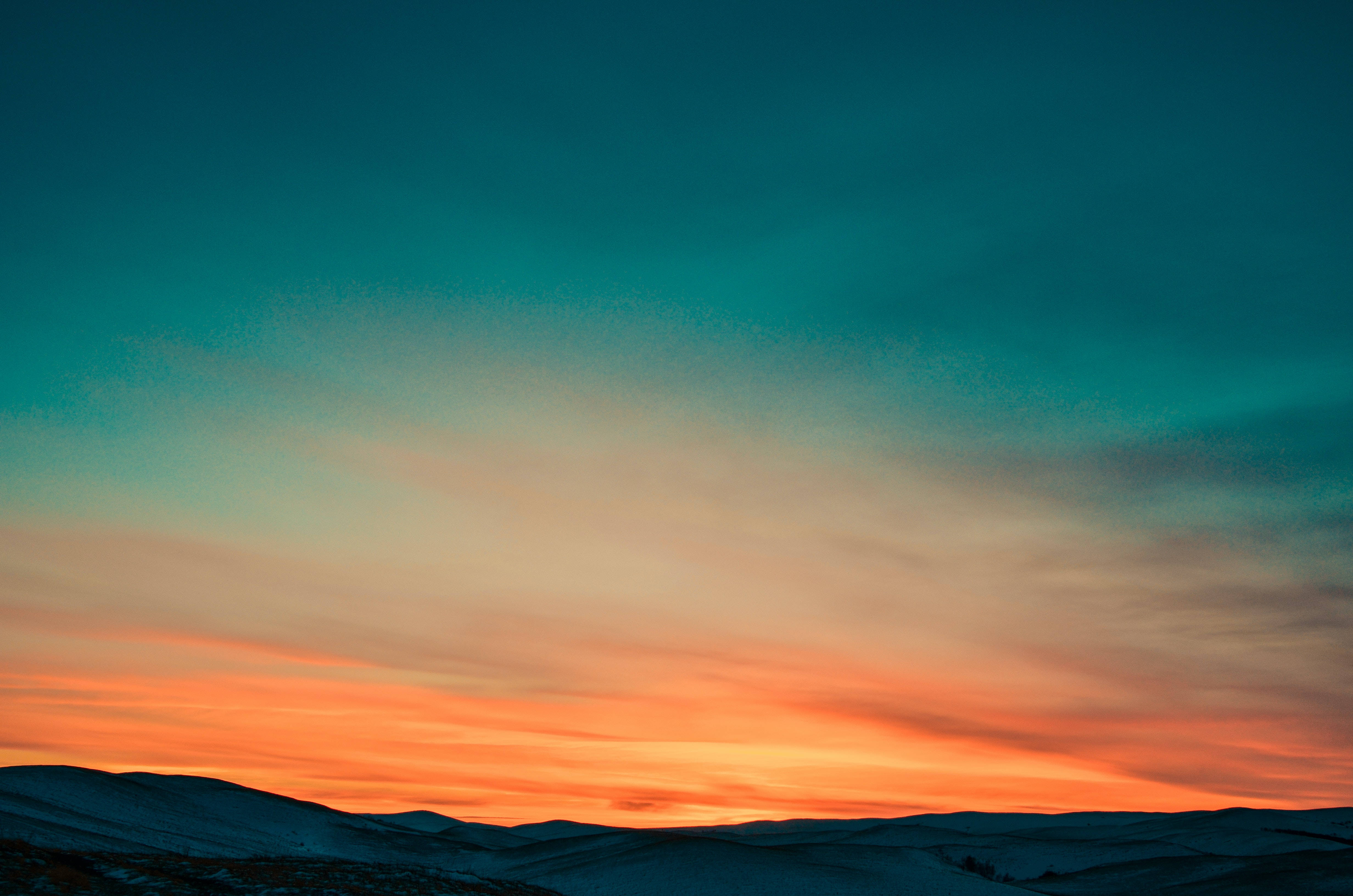 青橙交織的唯美天空 壁紙 — 白雪覆蓋的山脈在夕陽映照下形成剪影，天空融合了橙色和藍綠色的鮮豔色調。