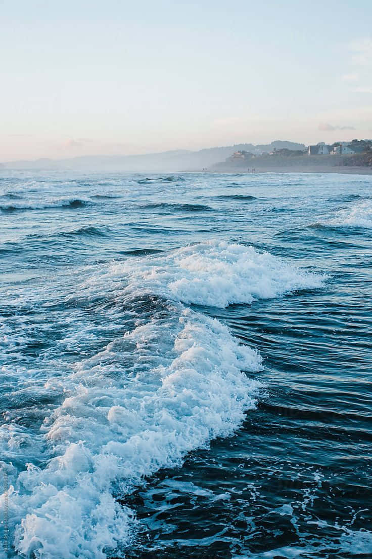 青綠色海景美學圖 壁紙 — 泡沫海浪在柔和粉彩天空下，伴隨著遠處海岸山丘和建築物，向岸邊翻滾。