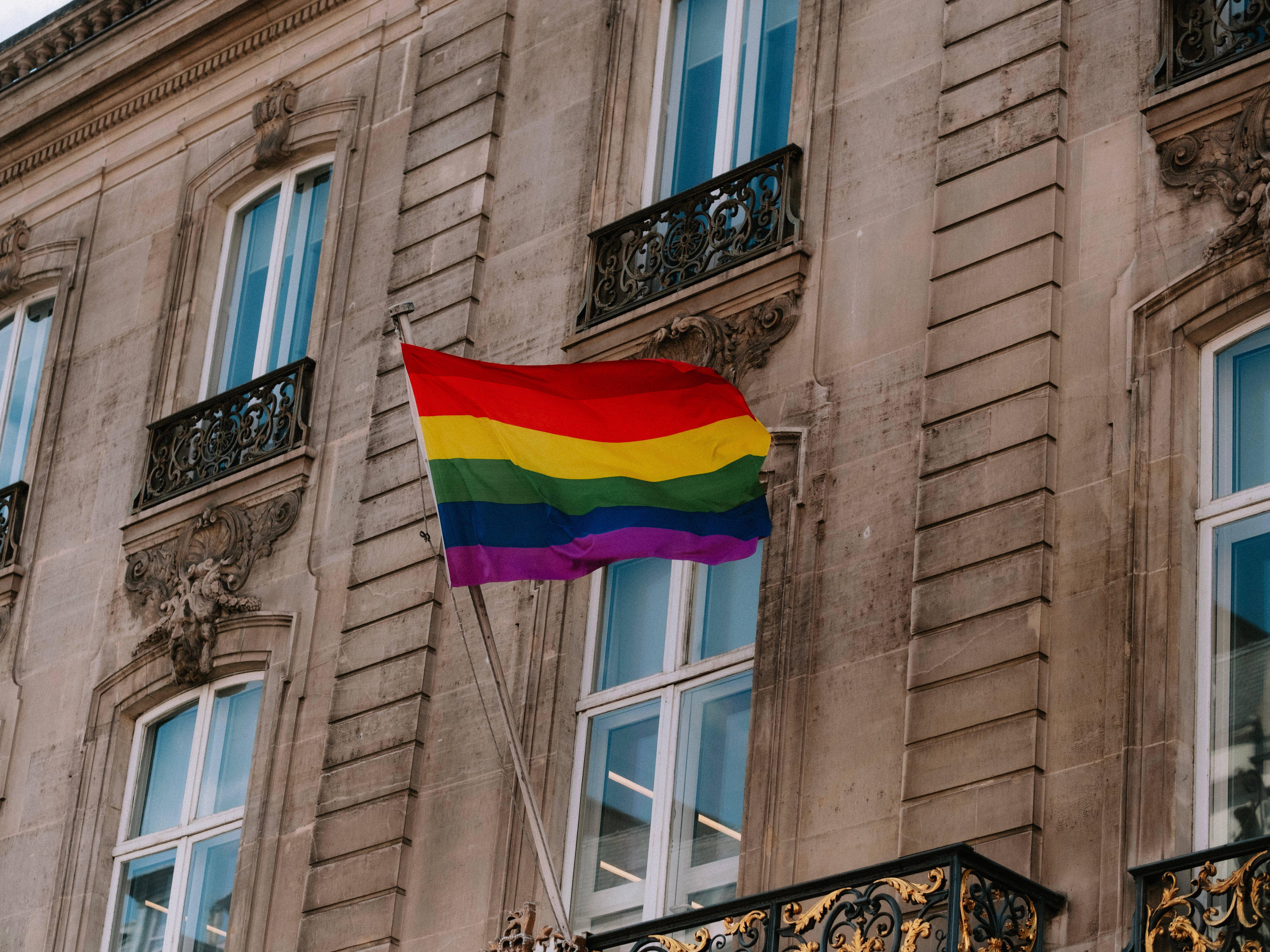 非二元旗幟象徵LGBT+社群 壁紙 — 一面鮮豔的彩虹旗在歷史悠久的石建築前自豪地飄揚，建築物有華麗的陽台和高窗。