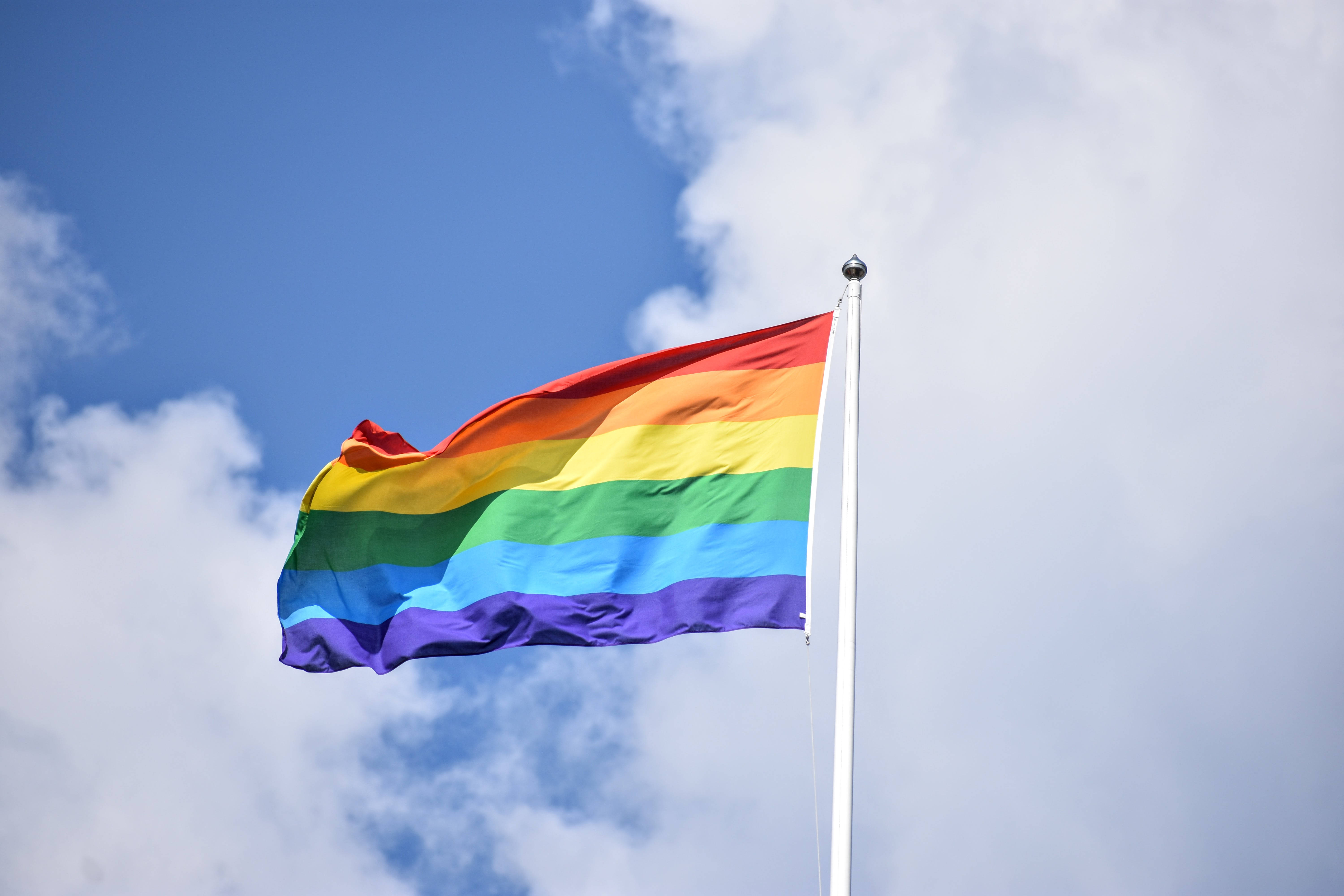 非二元與LGBT旗幟 壁紙 — 一面鮮豔的彩虹旗在藍天白雲下驕傲地飄揚在旗桿上。