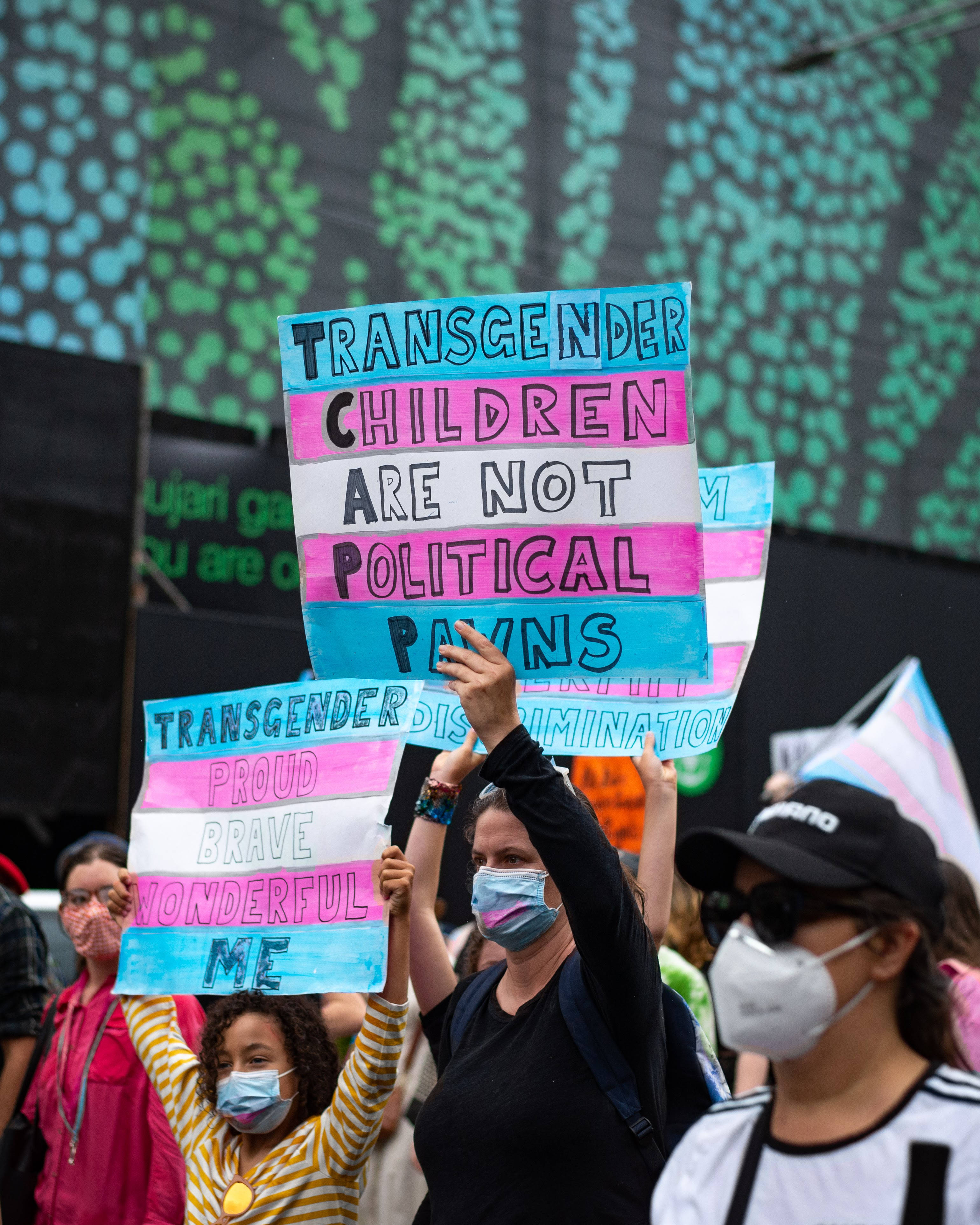 非二元與跨性別兒童 壁紙 — 抗議者在日間集會中高舉鮮豔的彩虹標語，倡導跨性別兒童的權益，背景帶有圖案。